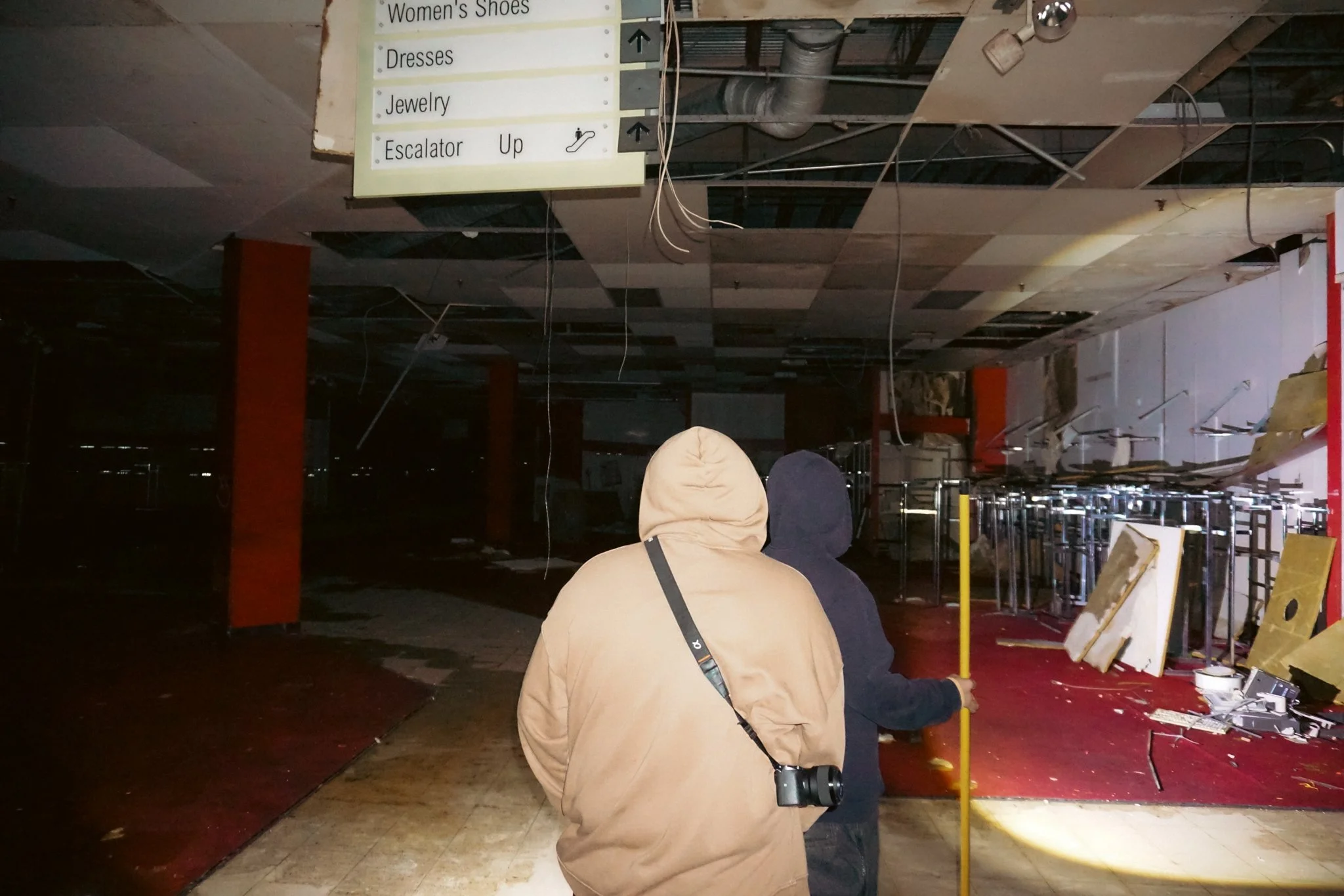 Two people in hooded jackets, one with a camera around their neck, walk through a damaged, abandoned shopping mall. The ceiling is partially collapsed with hanging wires, and debris is scattered on the floor. Signage points to women's shoes, dresses,