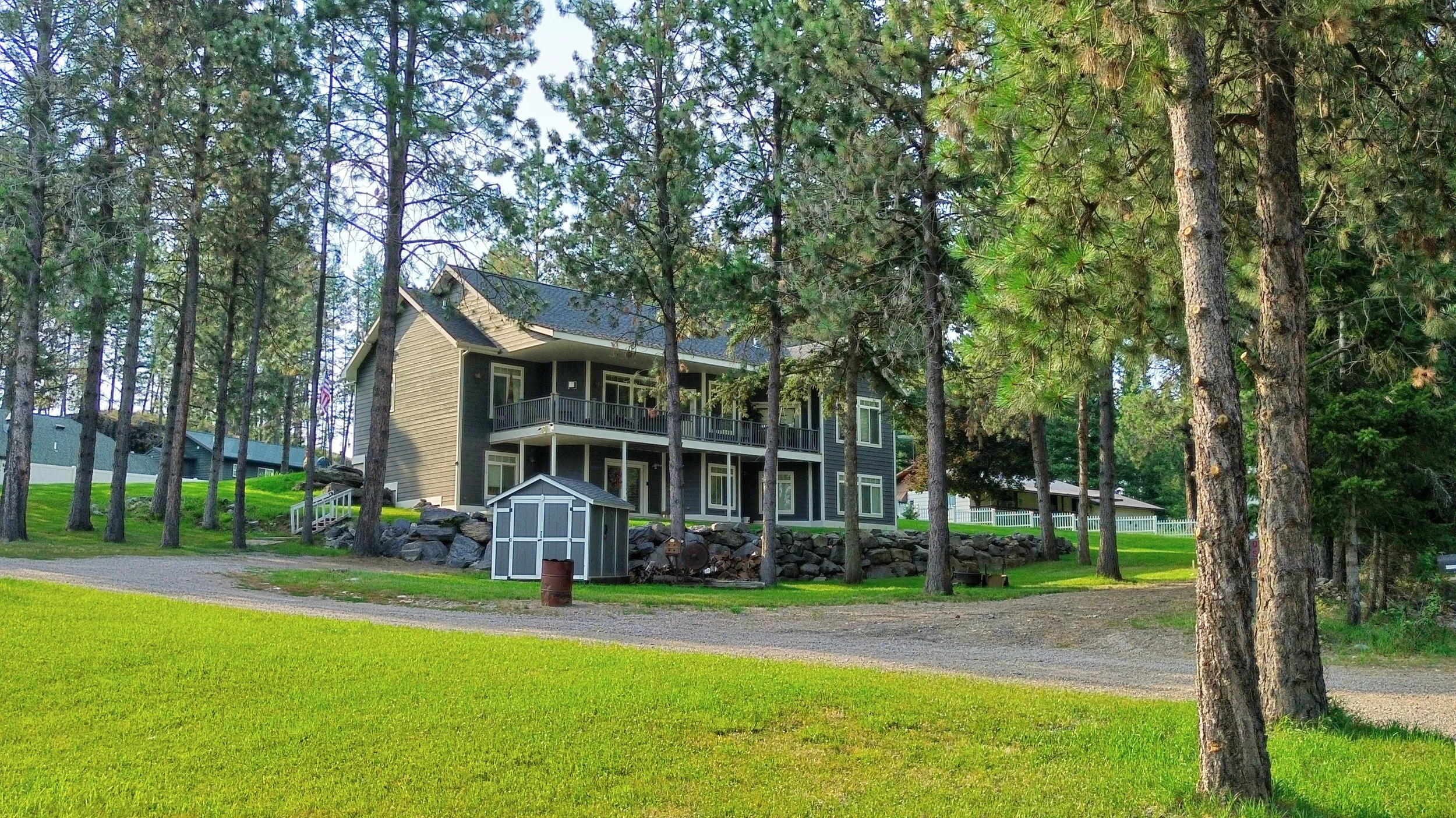 A two-story house painted gray with white trim, surrounded by tall pine trees, a well-maintained lawn, and a small storage shed in the front yard.