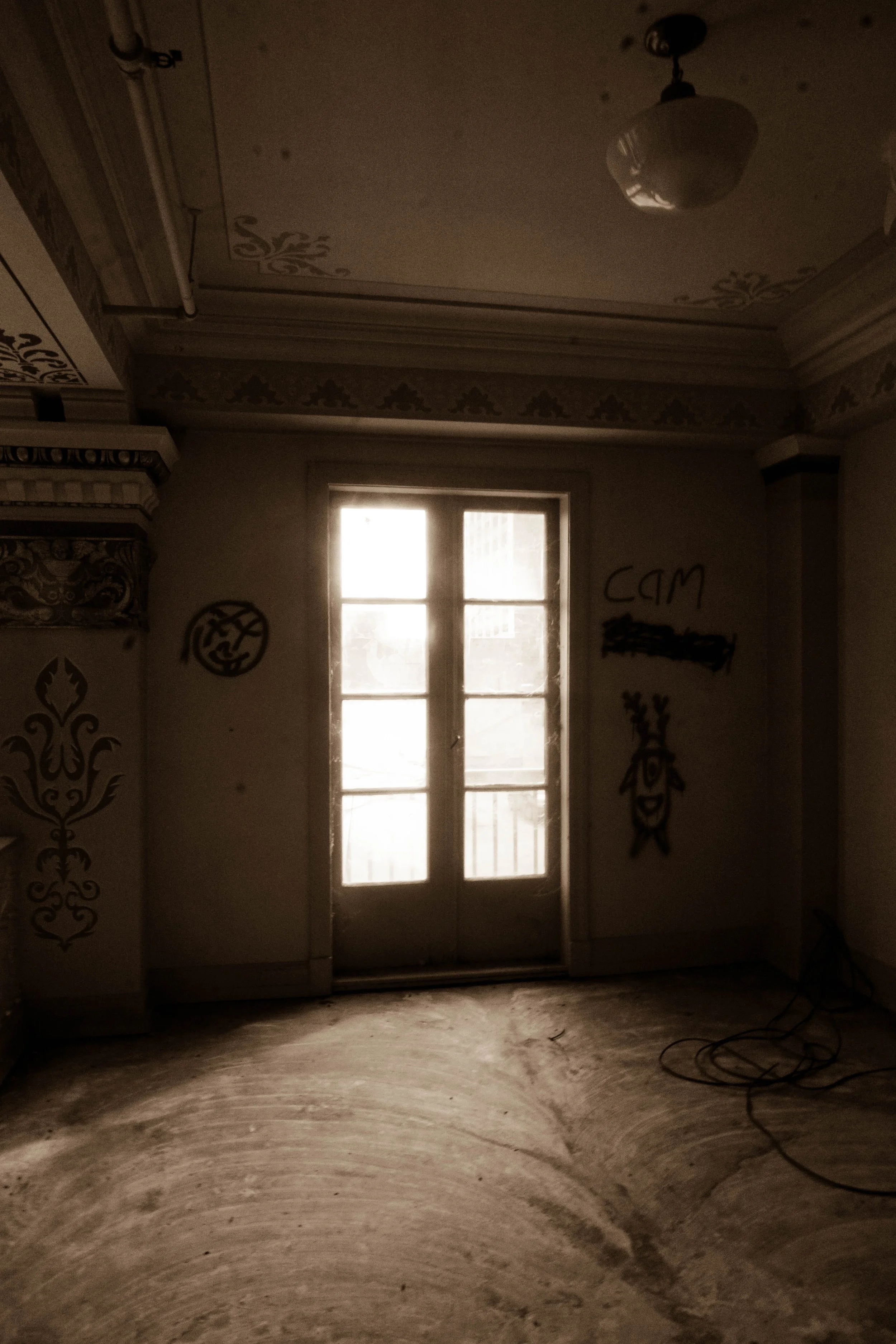 An interior room with a door that has glass panes, allowing light to enter. The room has decorative moldings on the ceiling and walls, and some graffiti on the walls, including a smiley face, the word 'cam' and a face with spiky hair. The floor appea