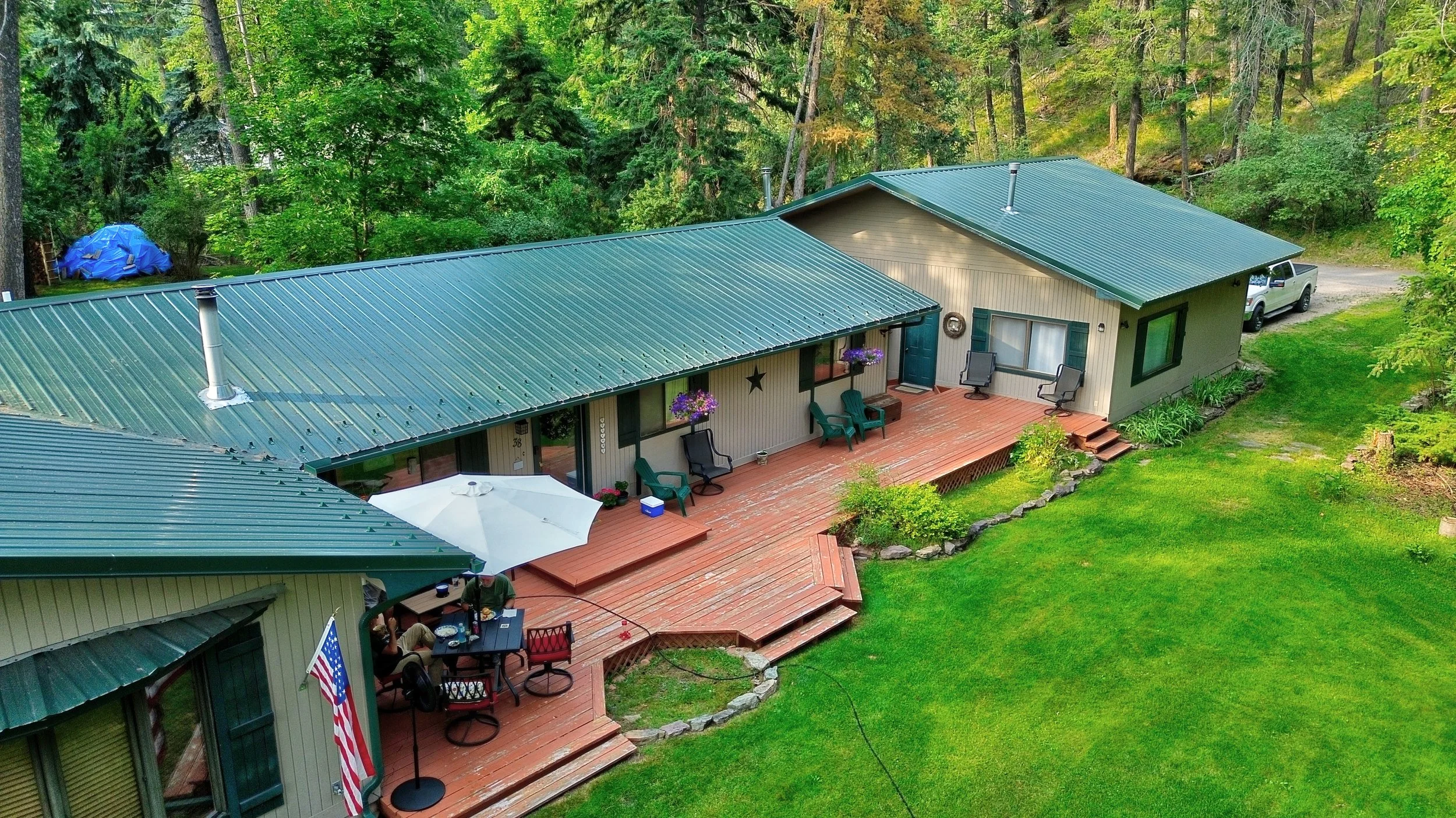 A house with green metal roofing, a wooden deck with outdoor furniture and purple flower pots, surrounded by a lush green yard and trees.