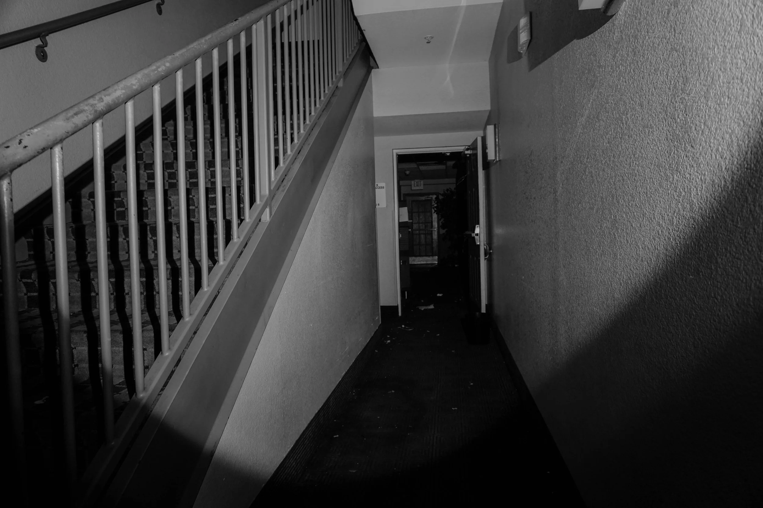 A dimly lit hallway with stairs on the left leading to an upper floor, and an open door at the end of the hallway revealing a door with a window and a sign that reads 'EXIT' inside a building.