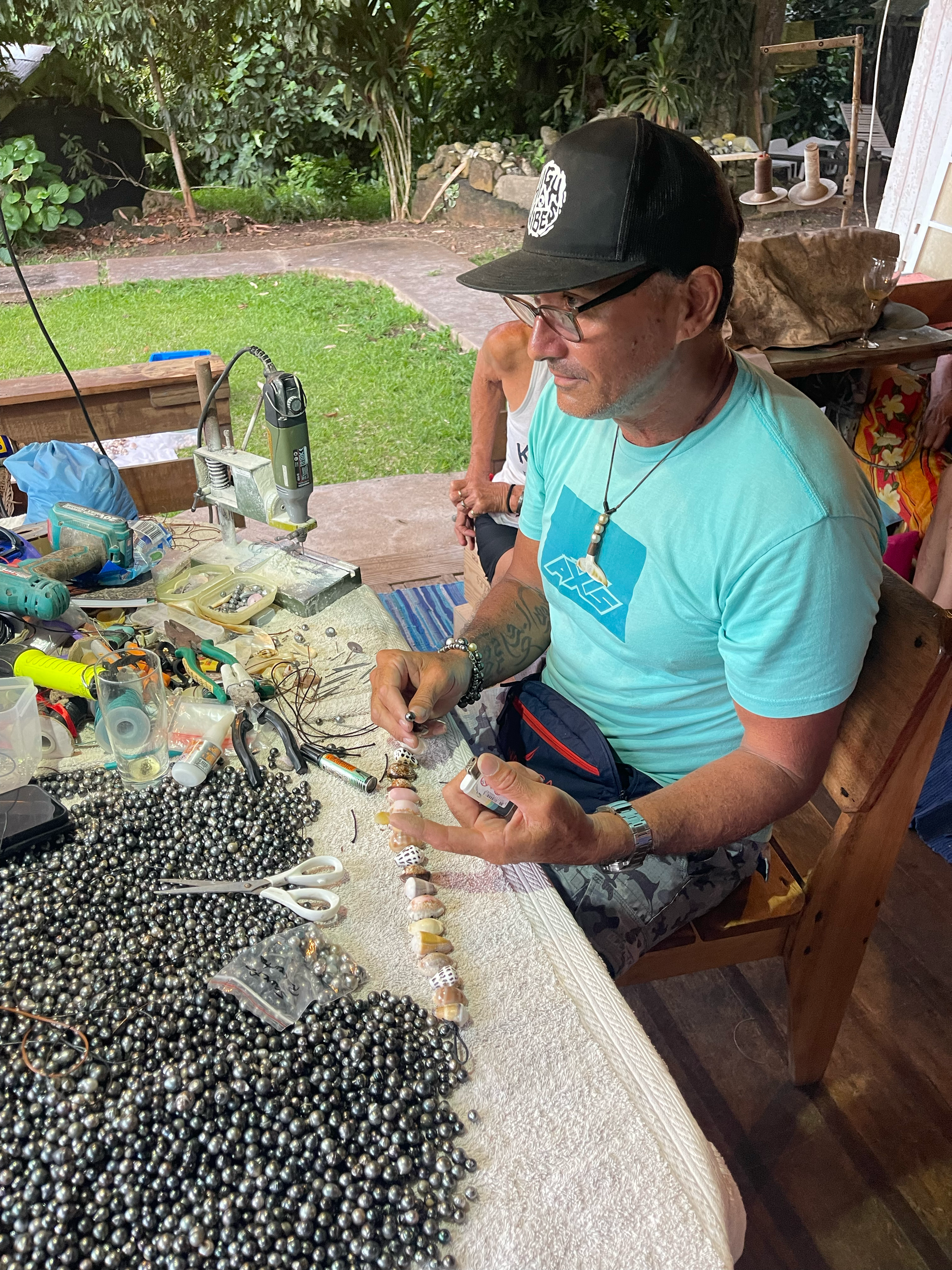 A man wearing a black cap, glasses, a light blue t-shirt, and camouflage shorts is sitting at a table with various jewelry-making supplies, including beads, tools, and elastic cords, outdoors amid greenery.
