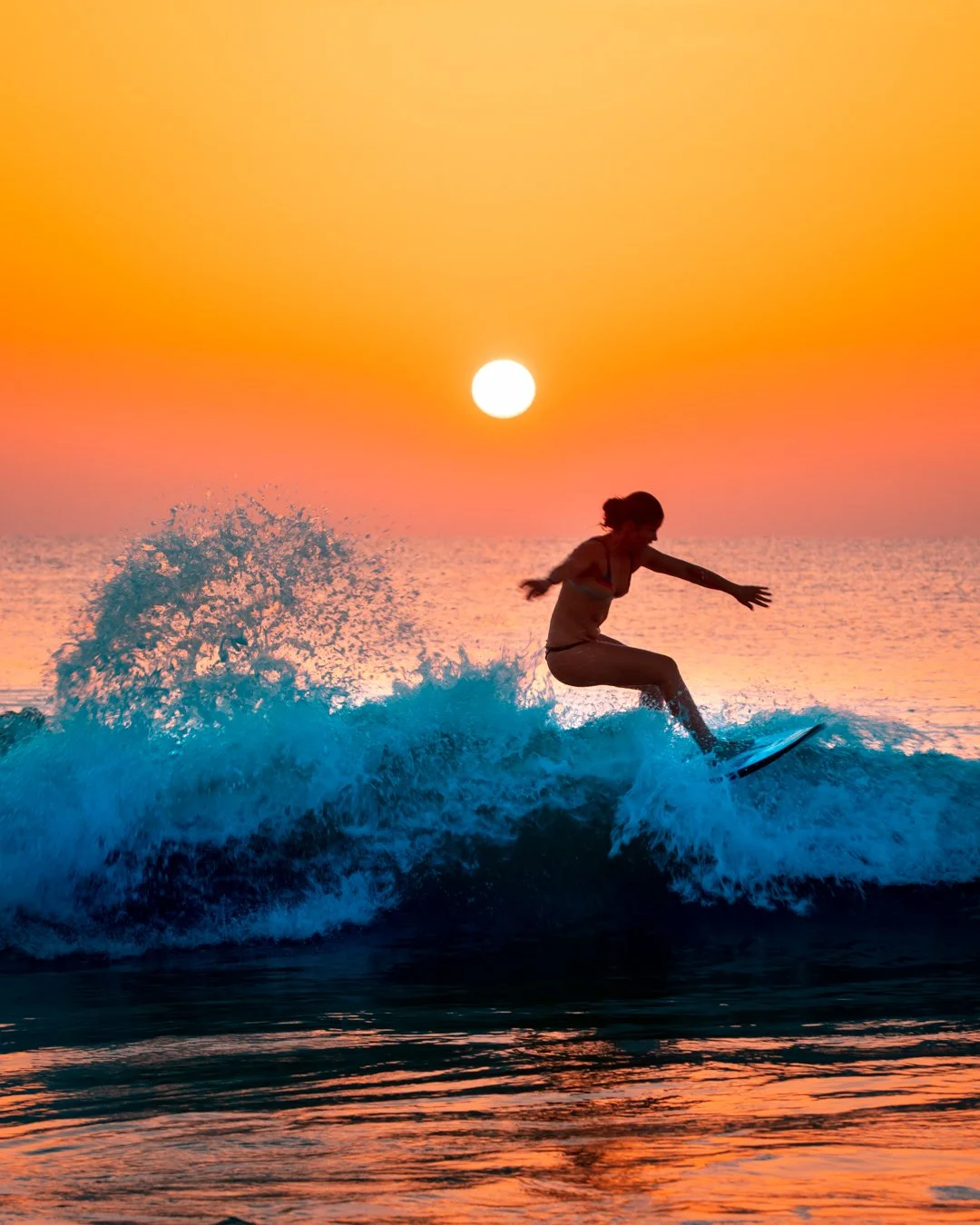 A woman surfing on a wave during sunset with an orange and pink sky and the sun near the horizon.