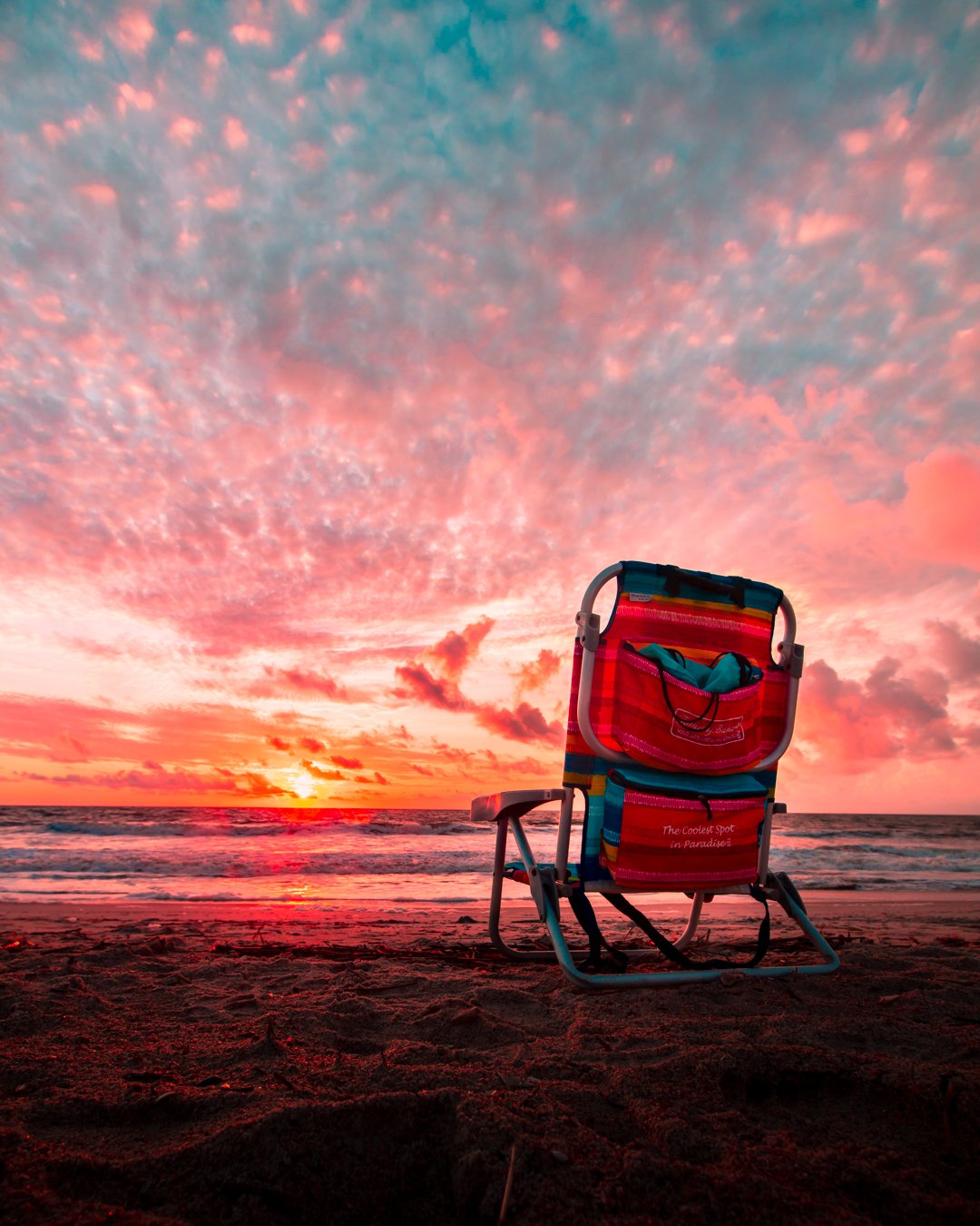 A colorful beach chair on the sand facing the ocean during a vibrant sunset with pink and orange sky.