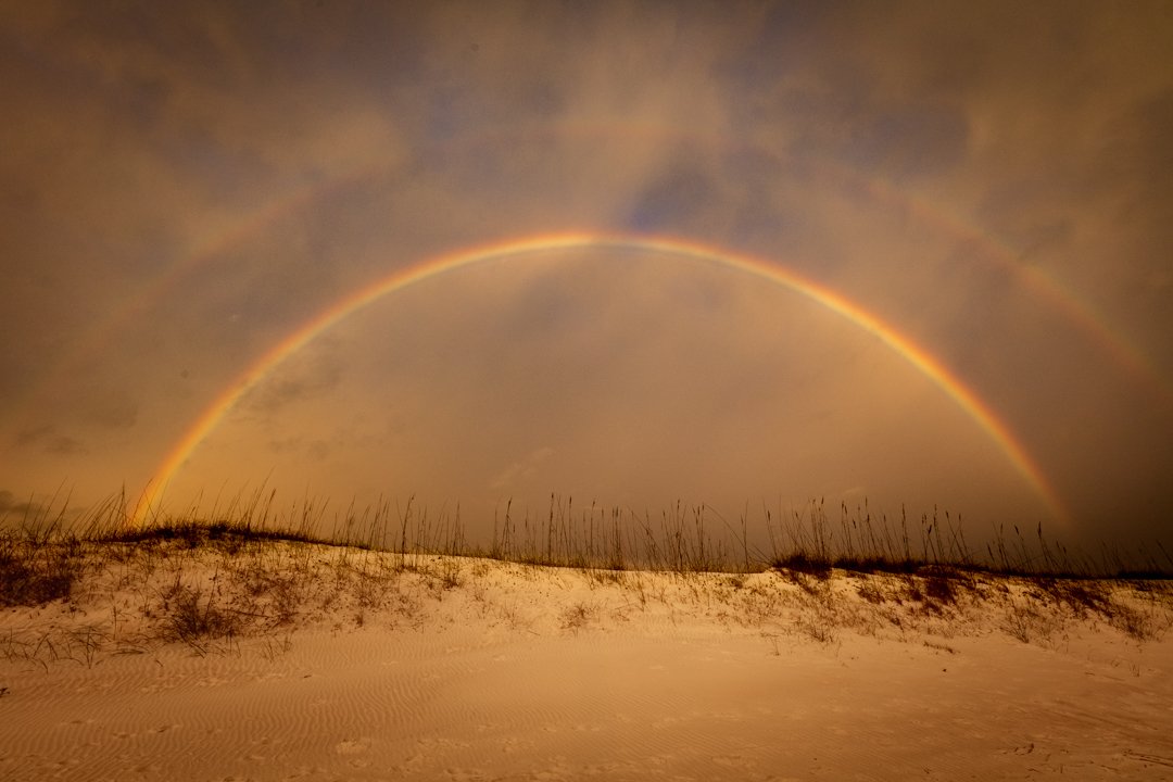 A double rainbow over a sandy beach with sparse grass.