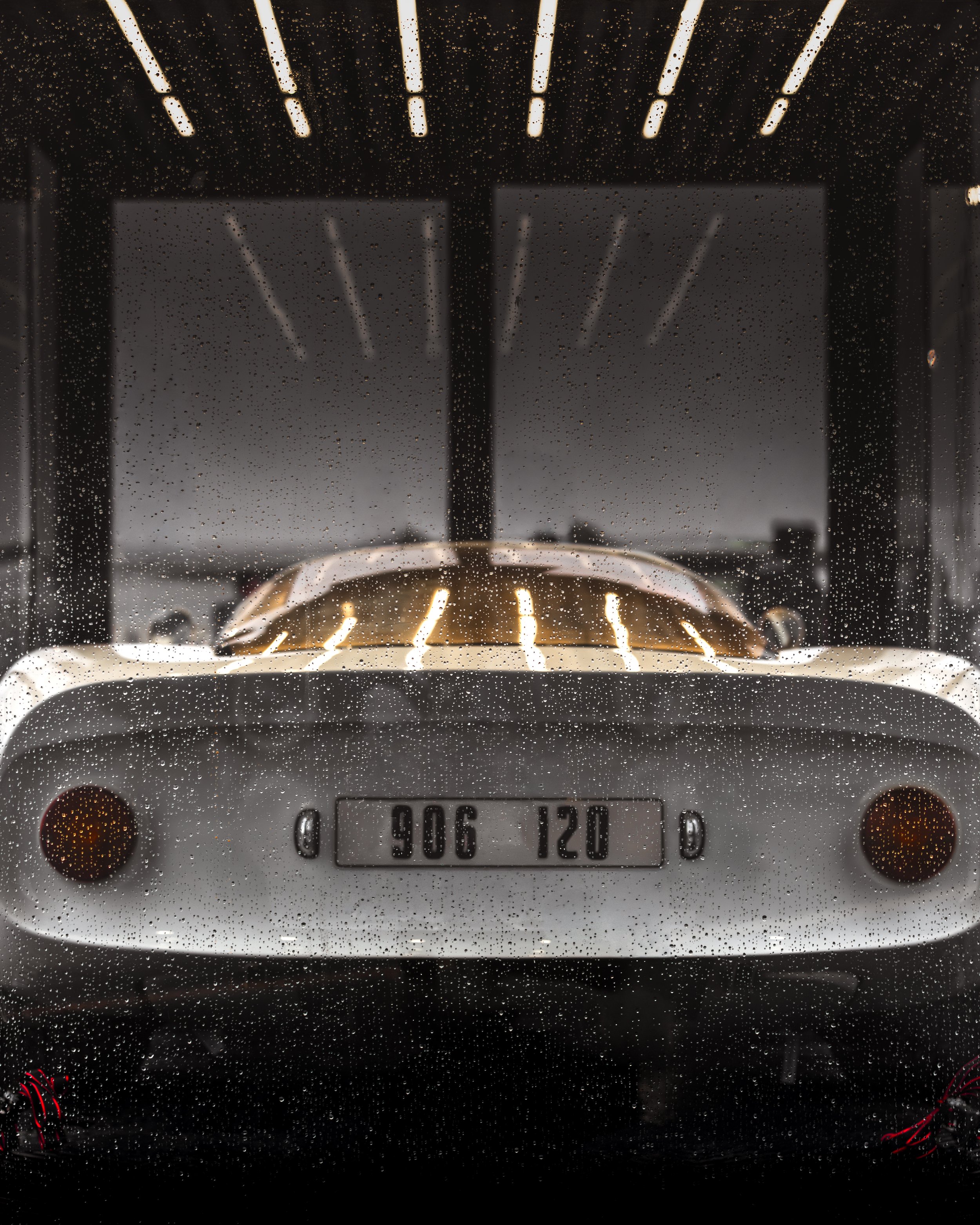 View of a classic white car seen through a rain-streaked window with raindrops on the glass. The car is a vintage model with round tail lights and a distinctive rear design.