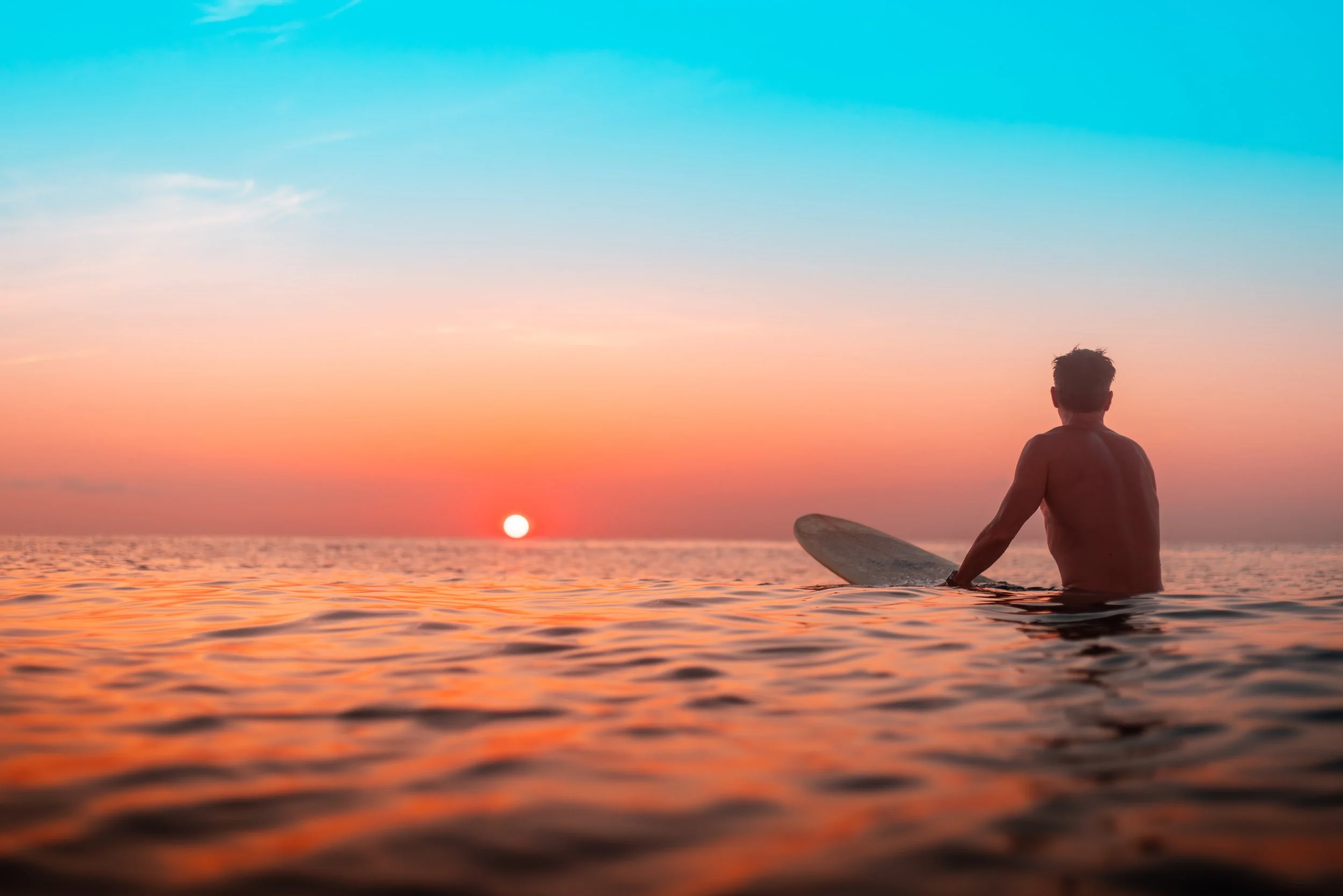 A person sitting on a surfboard in the ocean during a sunset, with the sun low on the horizon and the sky displaying shades of blue, pink, and orange.