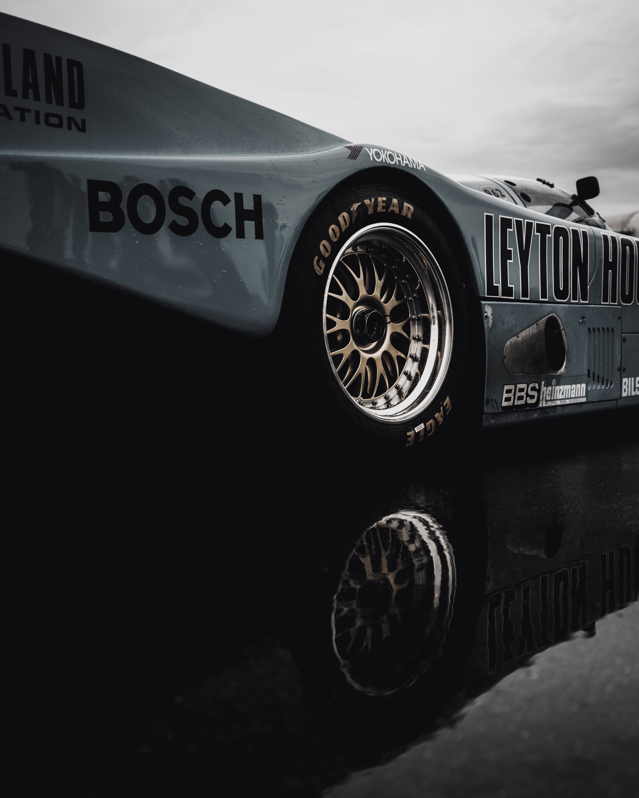 Close-up of a vintage race car with sponsor logos like Bosch, Leyton House, Yokohama, and Goodyear, reflected on a wet surface under an overcast sky.