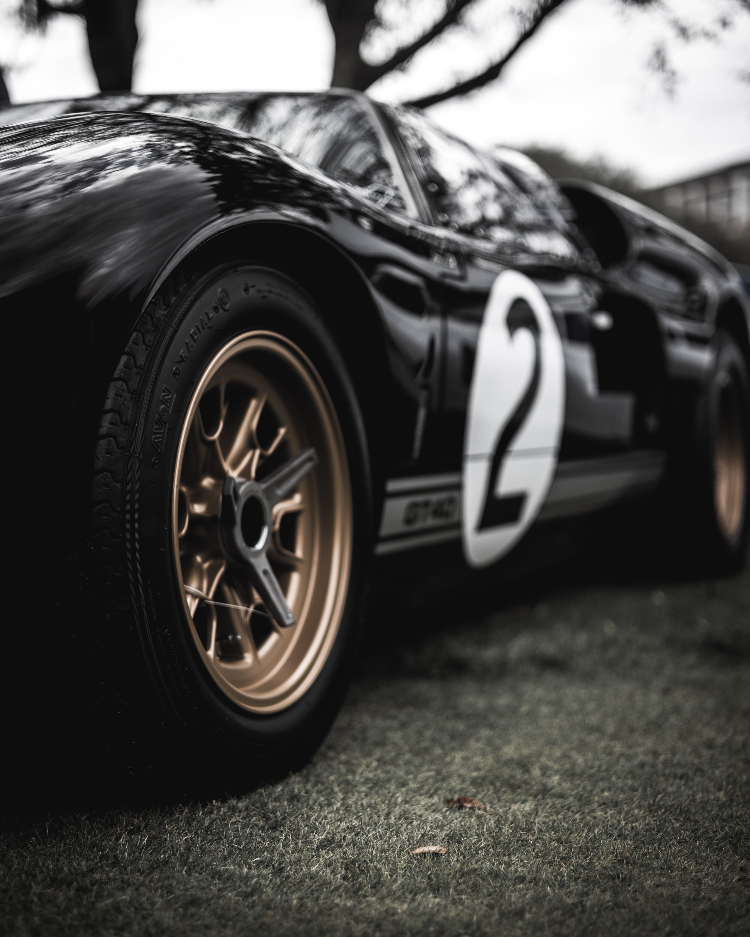 Close-up of a vintage black racing car with gold wheels, the number 2 displayed on the side, parked on grass with trees and a building in the background.