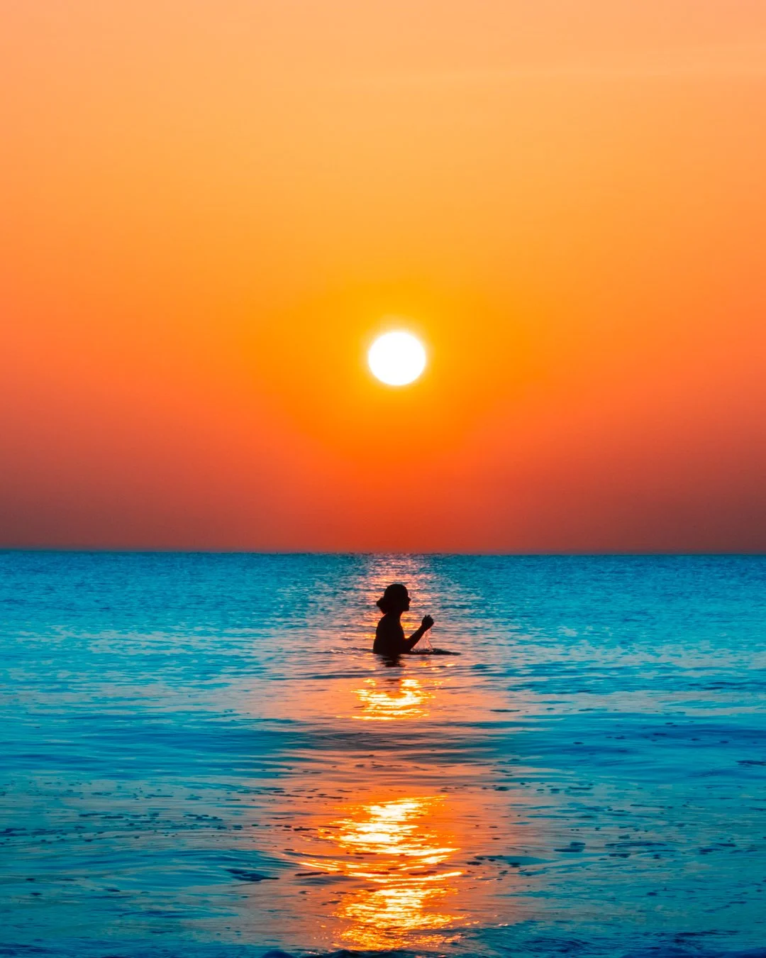 A person standing in the ocean during sunset, with the sun low on the horizon and colorful sky overhead.