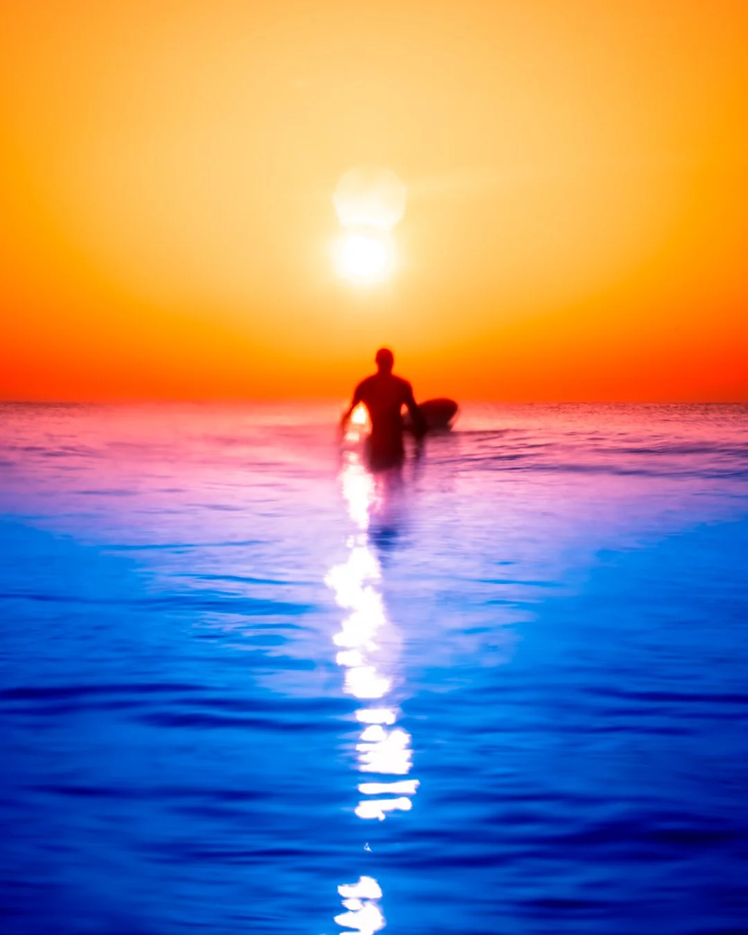 Silhouette of a person with a surfboard in the ocean at sunset, with vibrant orange, pink, and purple sky and a bright reflection on the water.