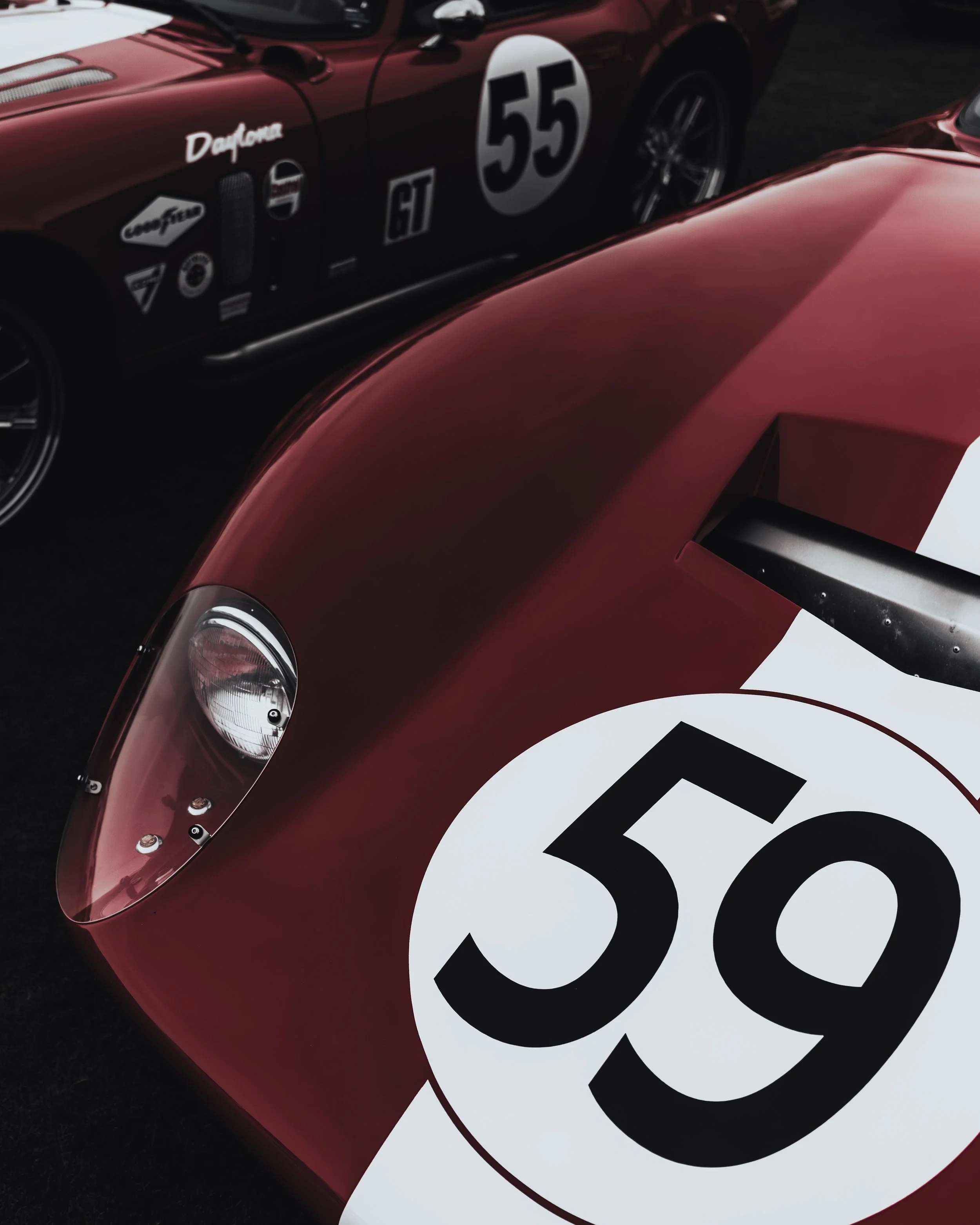 Close-up of vintage racing cars, with the front of a red car displaying a large white circle with the number 59 in black, and behind it a darker car with the number 55 and various race decals.