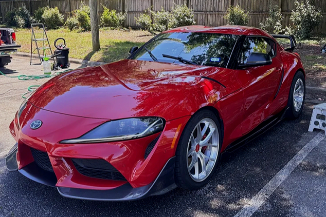 Red sports car parked on a driveway with a wooden fence and trees in the background. There are gardening tools and a ladder nearby.