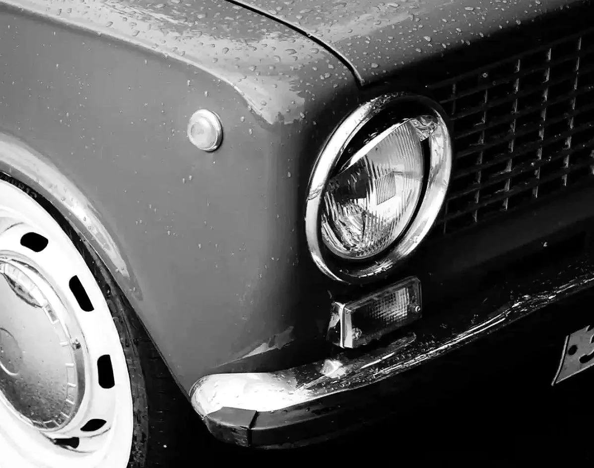 Close-up of the front left side of a vintage car with a round headlight, a small turn signal light, and a portion of the wheel and grille visible, all in black and white.