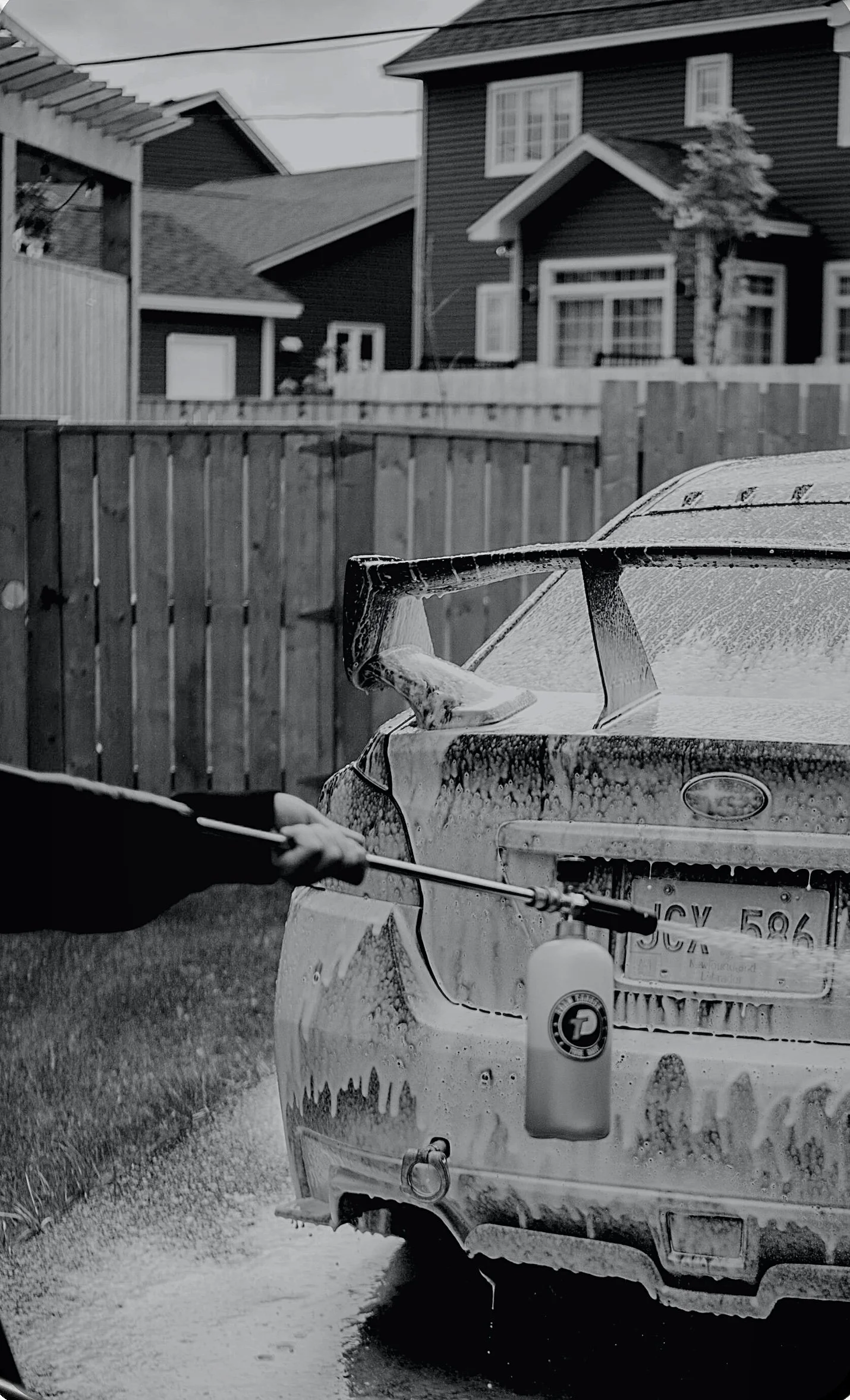 A person is washing a car with foam and a brush in a backyard, with houses and a wooden fence in the background.