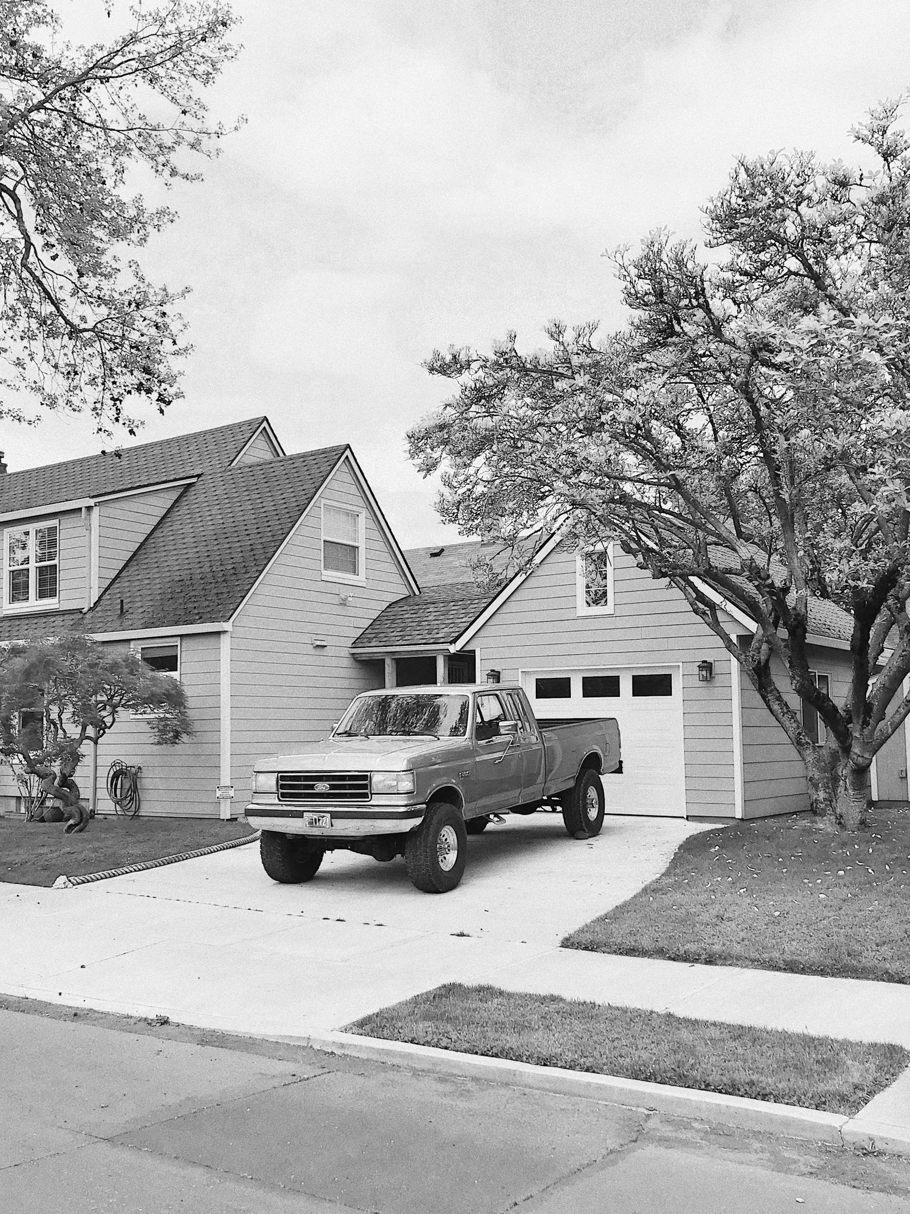 A residential house with a pickup truck parked in the driveway, some trees, and a garage with two small windows above it, in a monochrome image.