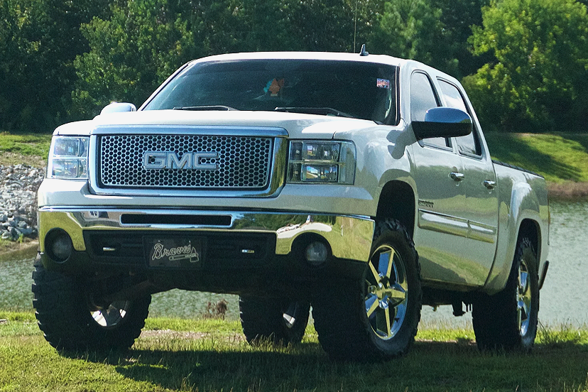 A white GMC pickup truck with large tires and chrome wheels parked on grass near a body of water with trees in the background.
