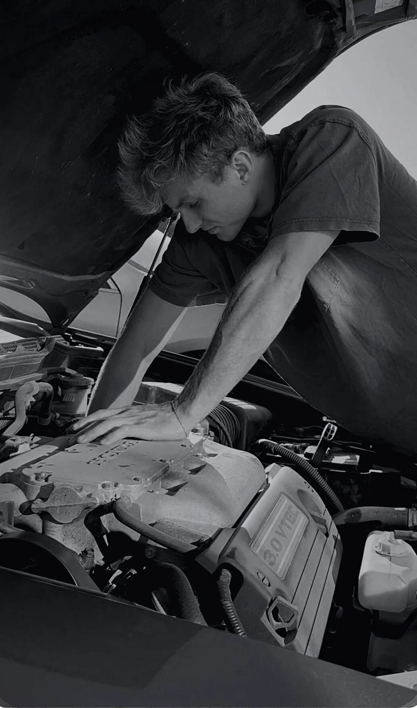 A person working under the hood of a car, inspecting or repairing the engine in a garage or workshop.