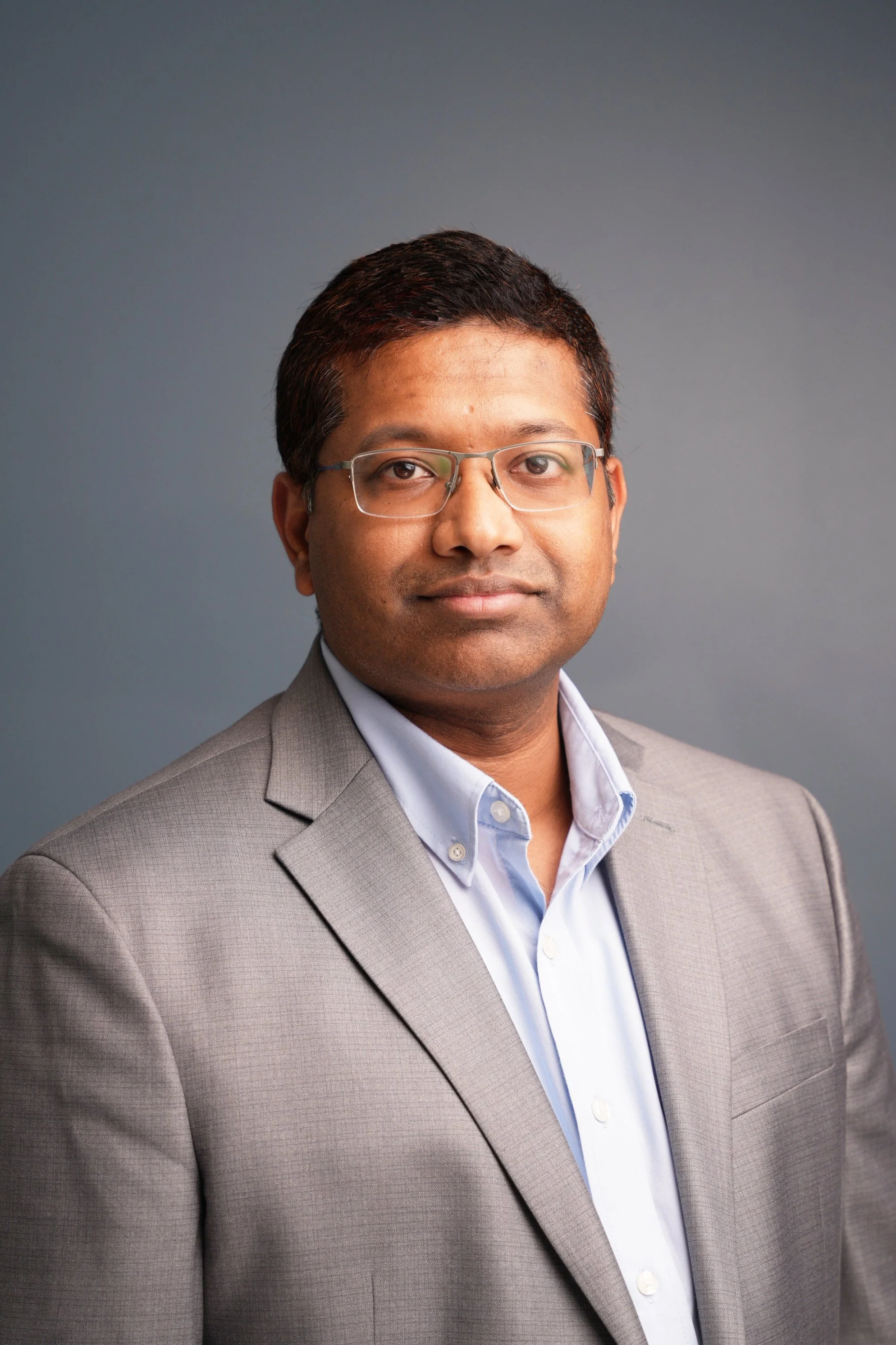 Professional headshot of a man in a grey suit, white shirt, and a striped maroon and grey tie, with short black hair, glasses, and a subtle smile against a plain light background.