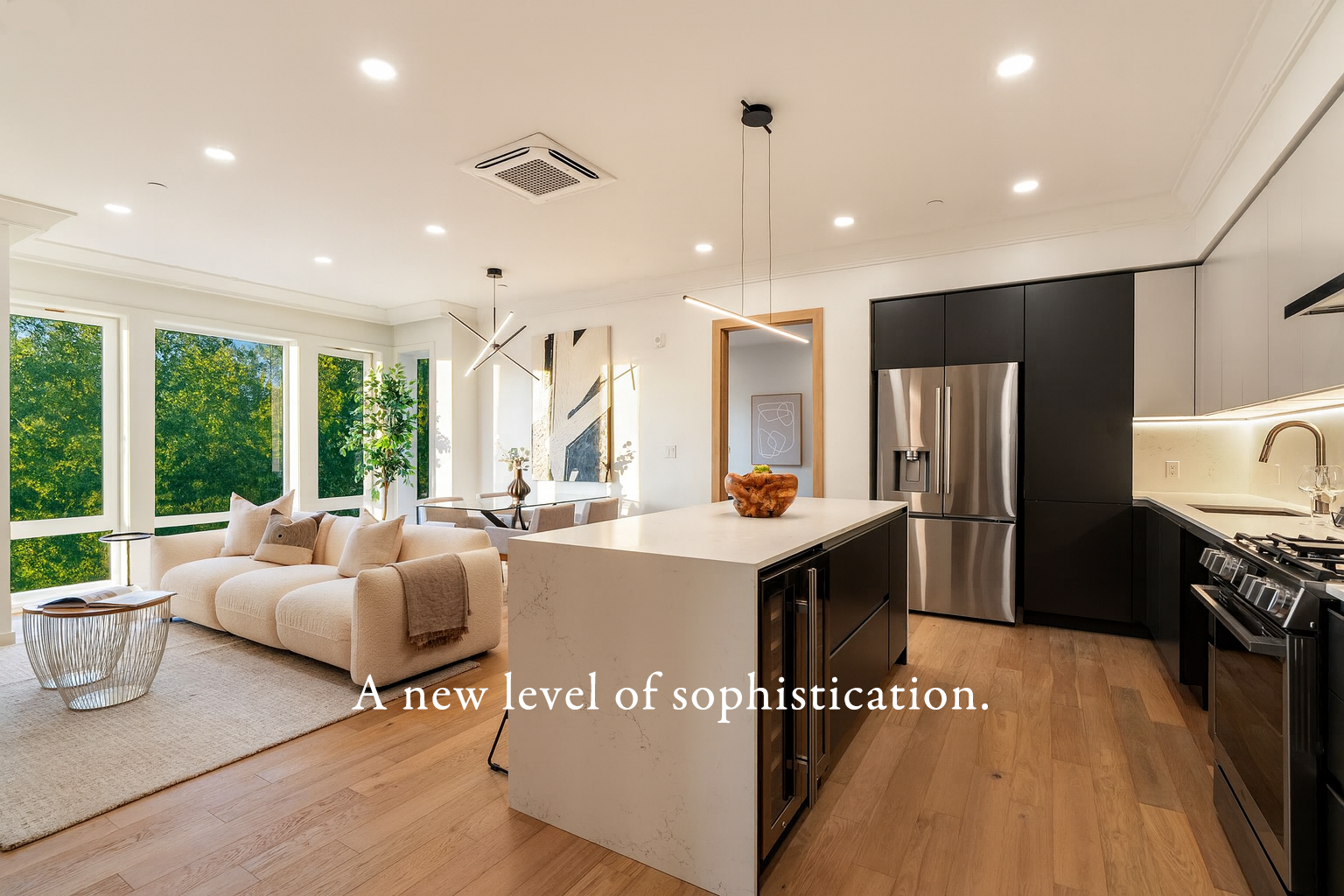 Modern open-concept living room and kitchen with large windows, beige sofa, abstract artwork, black and stainless steel appliances, white countertop, wooden flooring, and minimalistic decor.