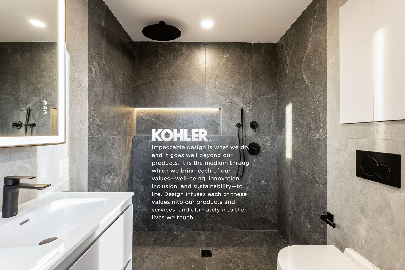 Modern bathroom with dark grey marble walls, white vanity with black faucet, large mirror, and a walk-in shower with black fixtures and overhead rain showerhead, Kohler branding in the center.