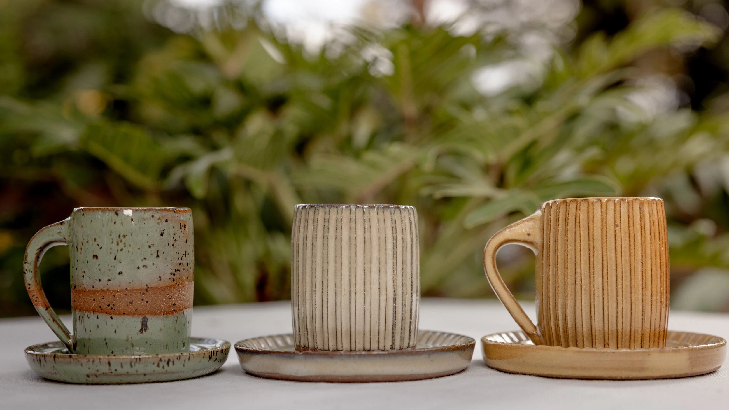 Three ceramic mugs with matching saucers, each with a different design and color, arranged in a row on a white surface outdoors. The background is blurred green foliage.