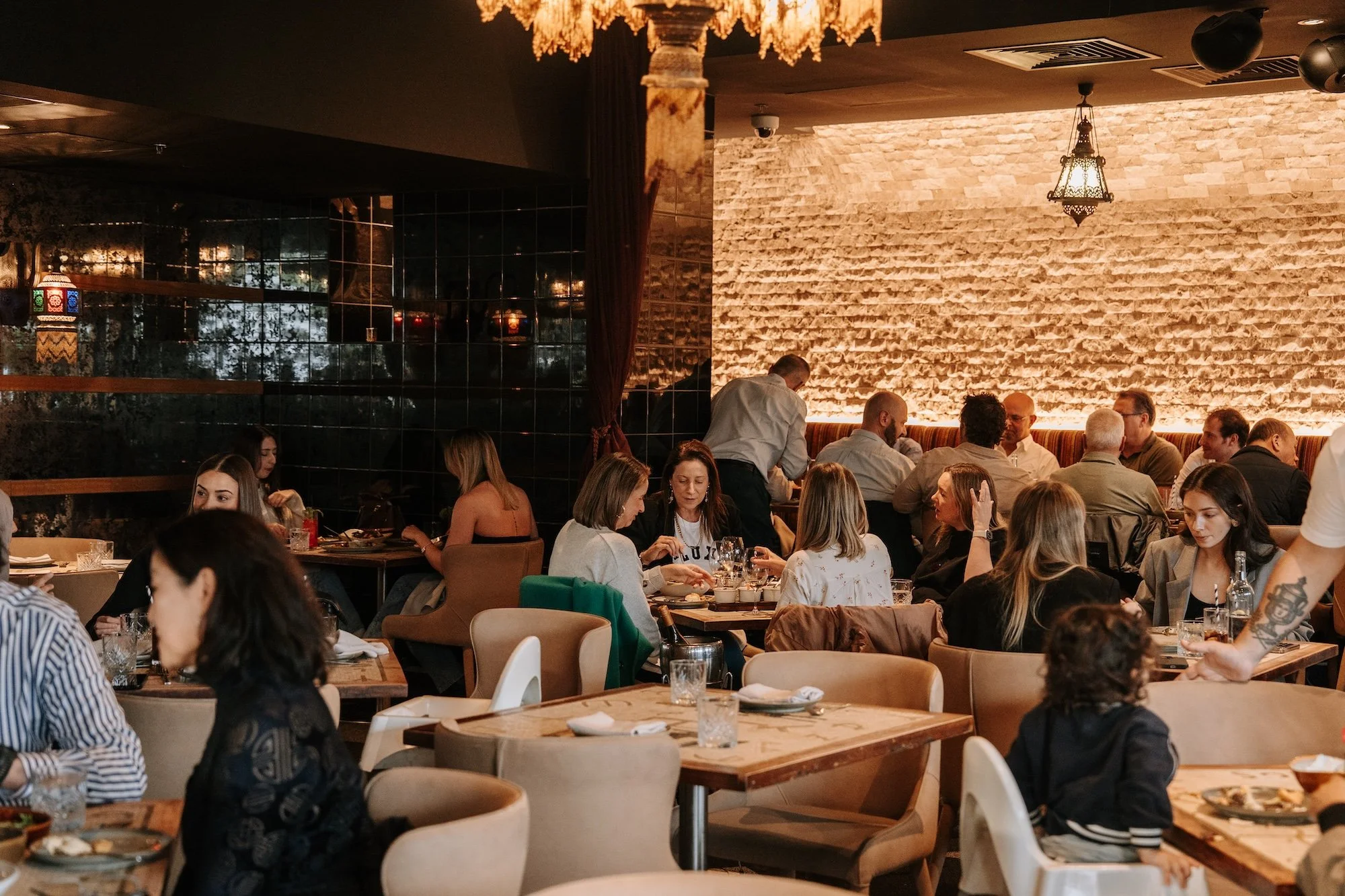 People dining and socializing inside a restaurant with warm lighting, brick wall, and modern decor.