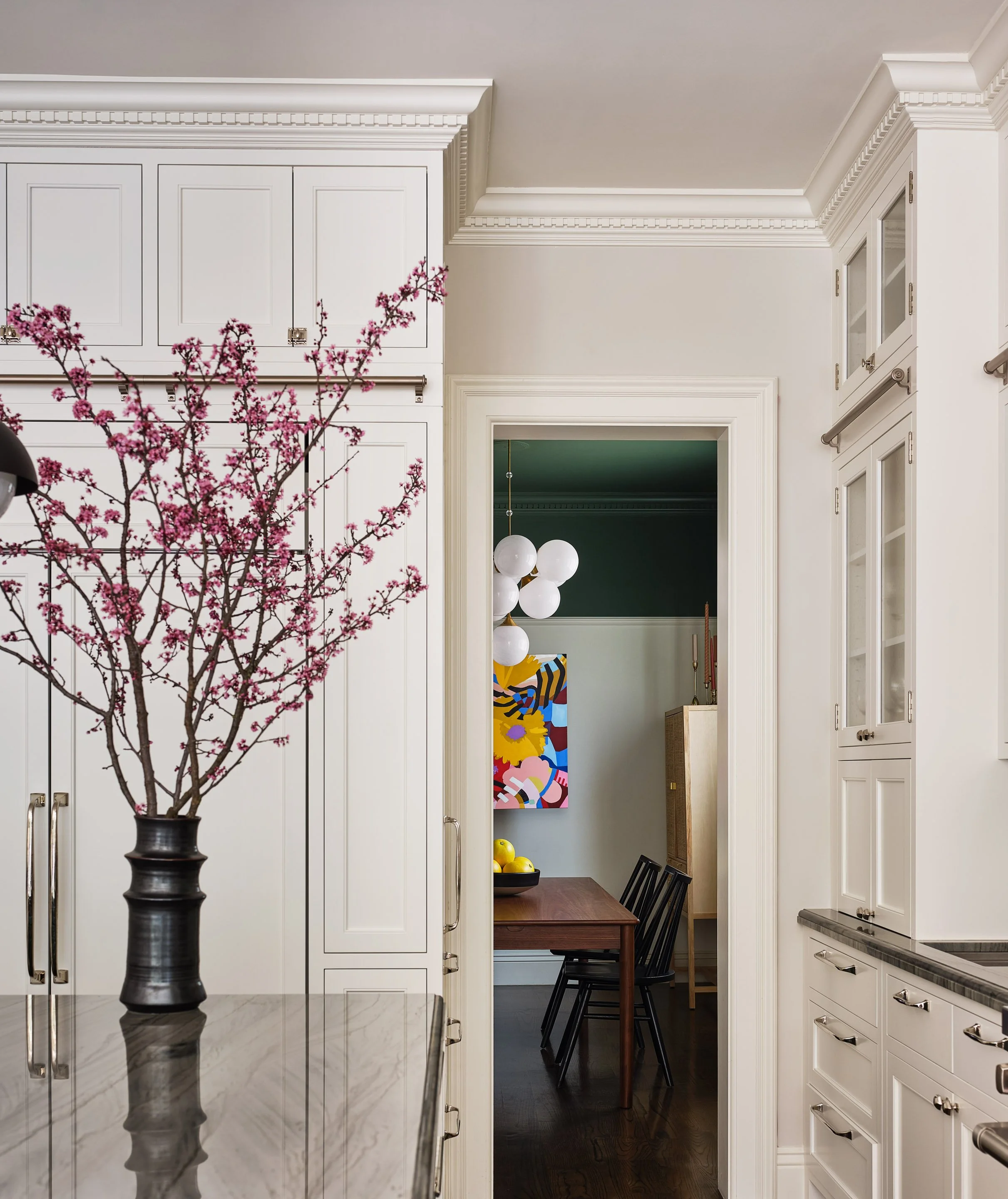 A white kitchen with a marble countertop, white cabinets, a black vase with pink blossoms, and a doorway leading into a dining area with a colorful painting and black chairs.