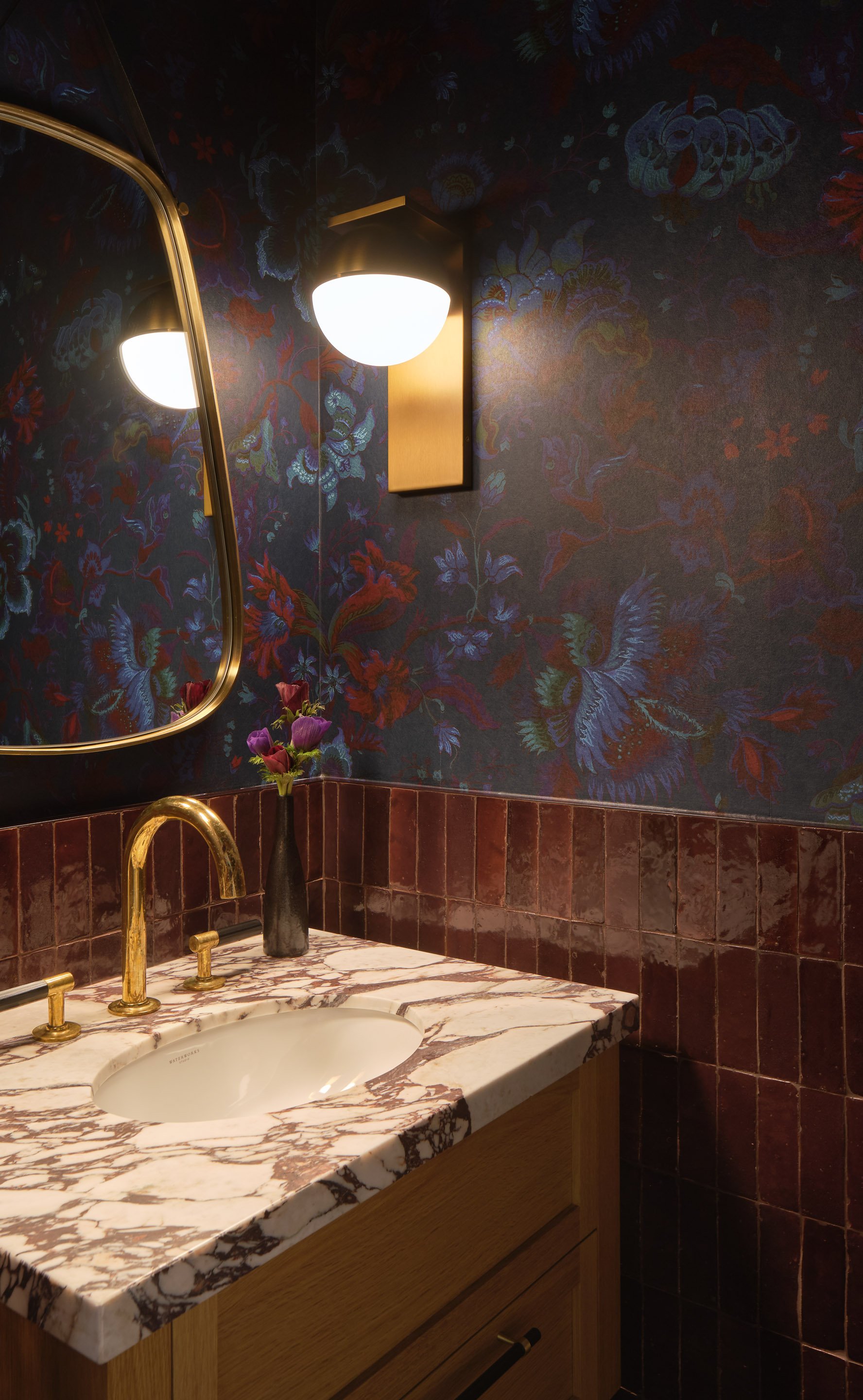 ACD | Alicia Cheung Design detailed this powder room with jewel-toned floral wallpaper, brass fixtures, and a curved brass mirror, creating a moody and glamorous vignette perfect for a bold first impression.