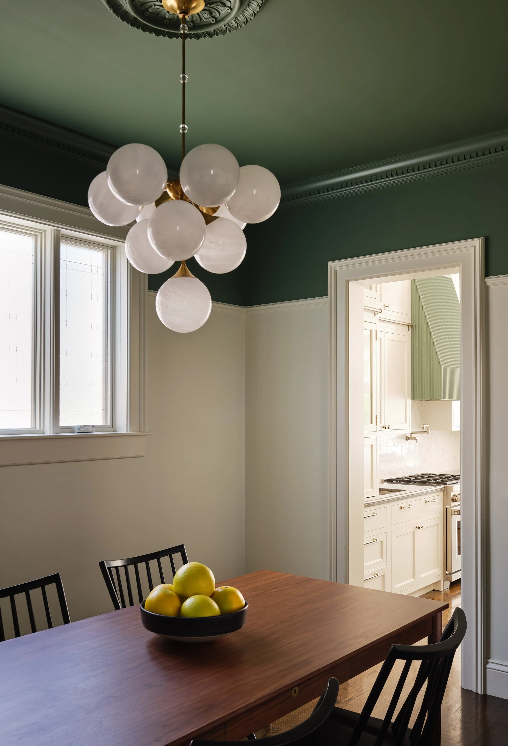 ACD | Alicia Cheung Design designed this dining room with a sculptural glass globe chandelier and a deep green ceiling, opening into a fluted sage green kitchen beyond, creating a layered and connected space perfect for entertaining.