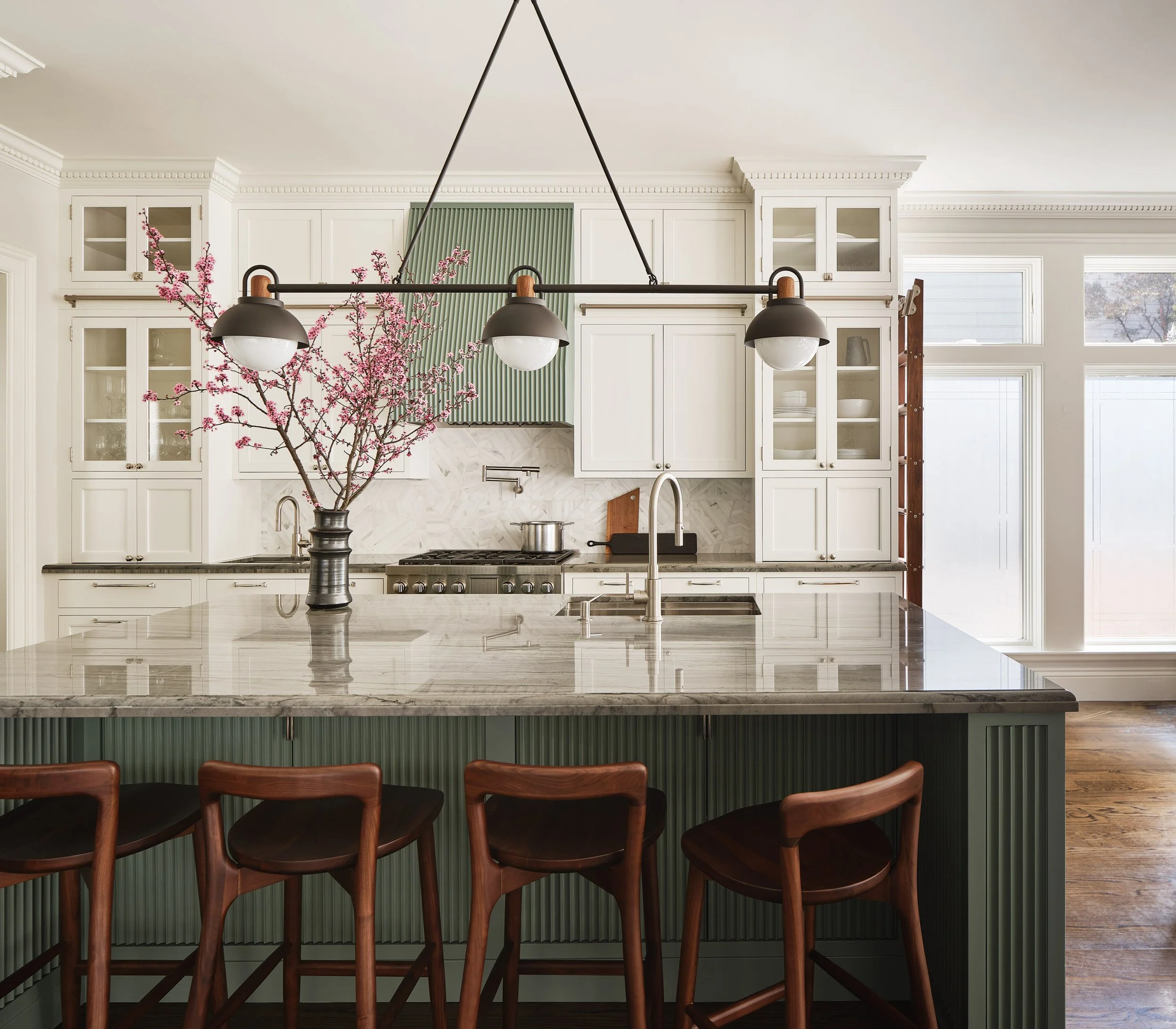 A modern kitchen with white cabinetry, a gray and white marble island, a black pendant light fixture, a branch with pink blossoms in a black vase on the island, and a green decorative panel on the wall.