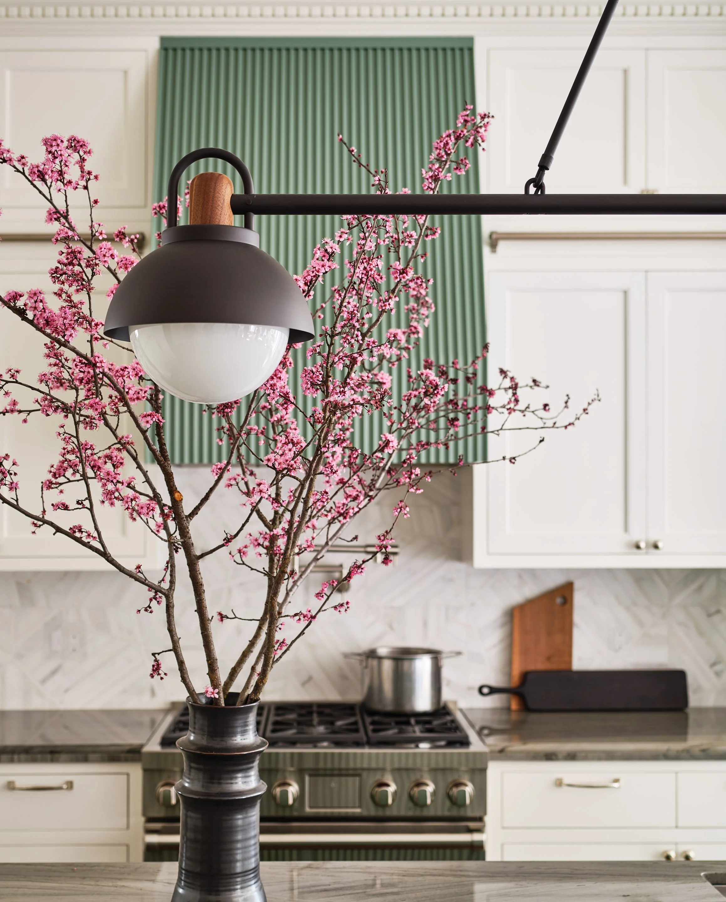 ACD | Alicia Cheung Design styled this kitchen vignette with flowering cherry branches, a globe pendant light, and a fluted sage green range hood, creating a warm and detailed moment perfect for everyday cooking.
