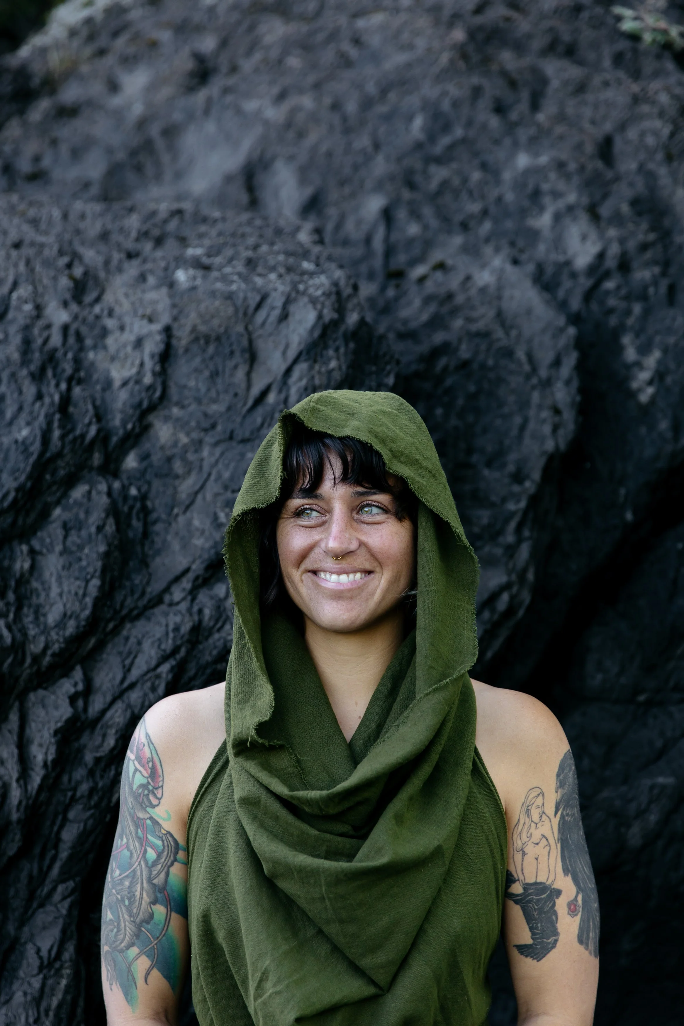 A smiling woman with tattoos on her arms, wearing a green hooded garment, standing in front of dark rocky terrain.