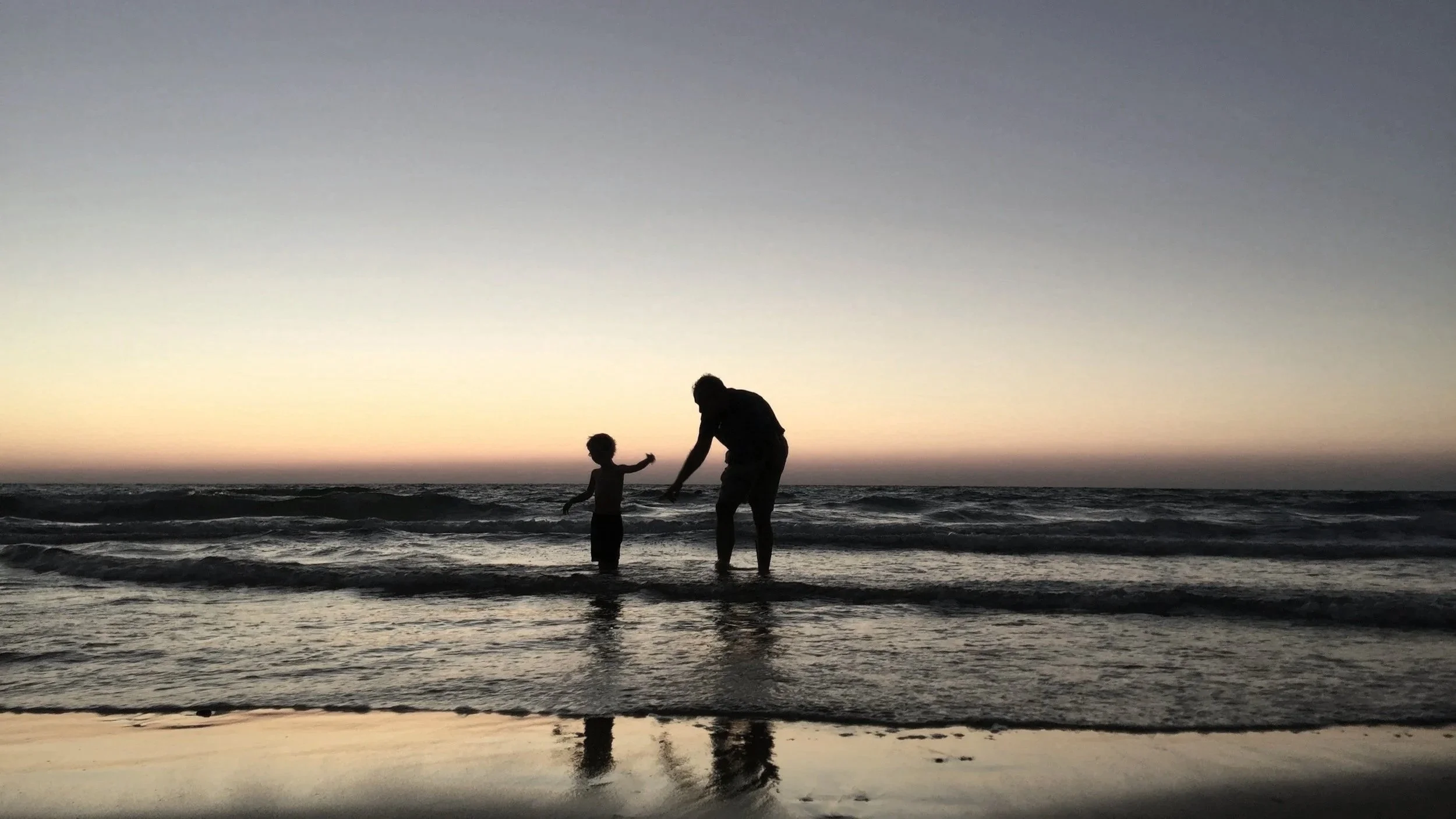 A silhouette of a parent and a child playing and holding hands on the beach during sunset.