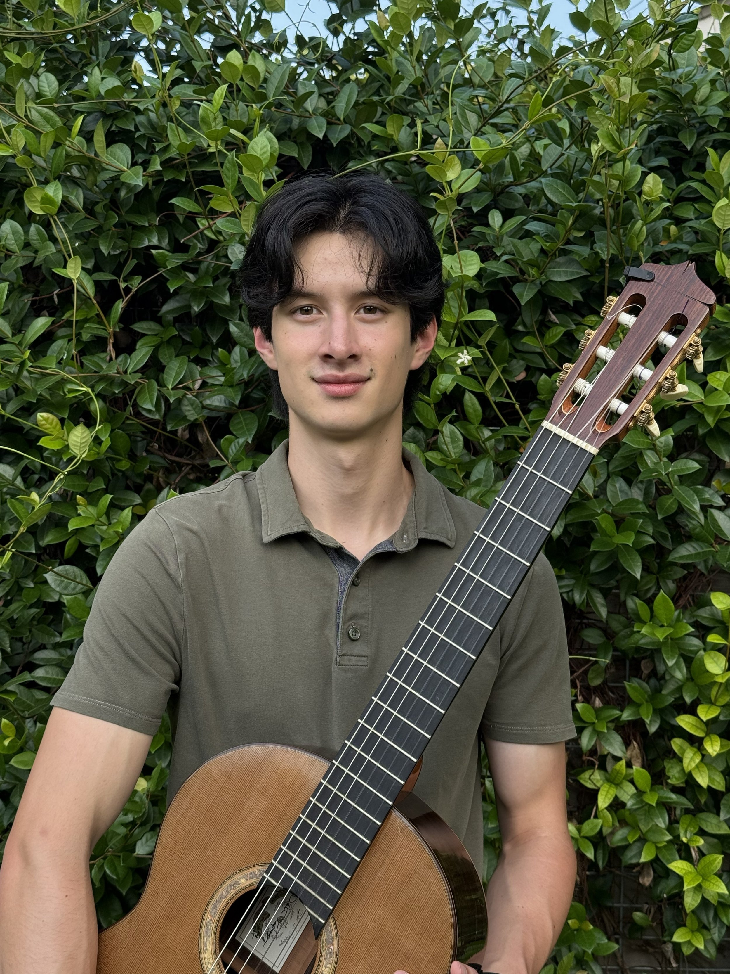 A young man with dark hair and a light olive green shirt holding a guitar in front of a backdrop of lush green leaves.