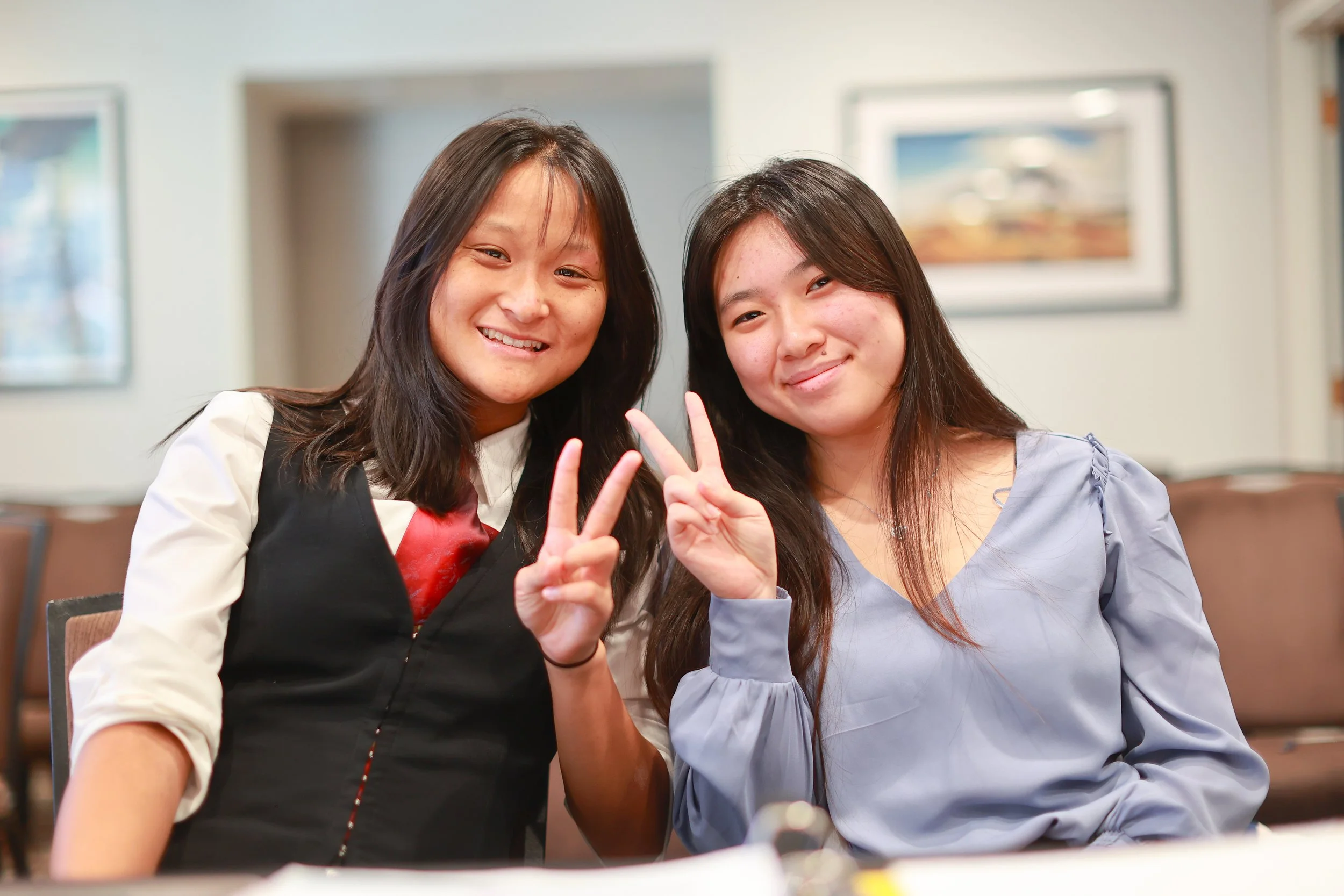 Two women sitting indoors, smiling and making peace signs with their hands.