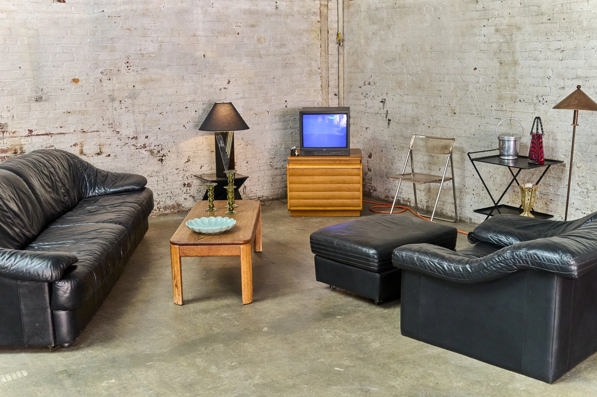Living room with black leather sofa, black leather armchair with ottoman, wooden coffee table with decorative objects, small TV on a wooden cabinet, and industrial decor elements, including a brick wall and concrete floor.