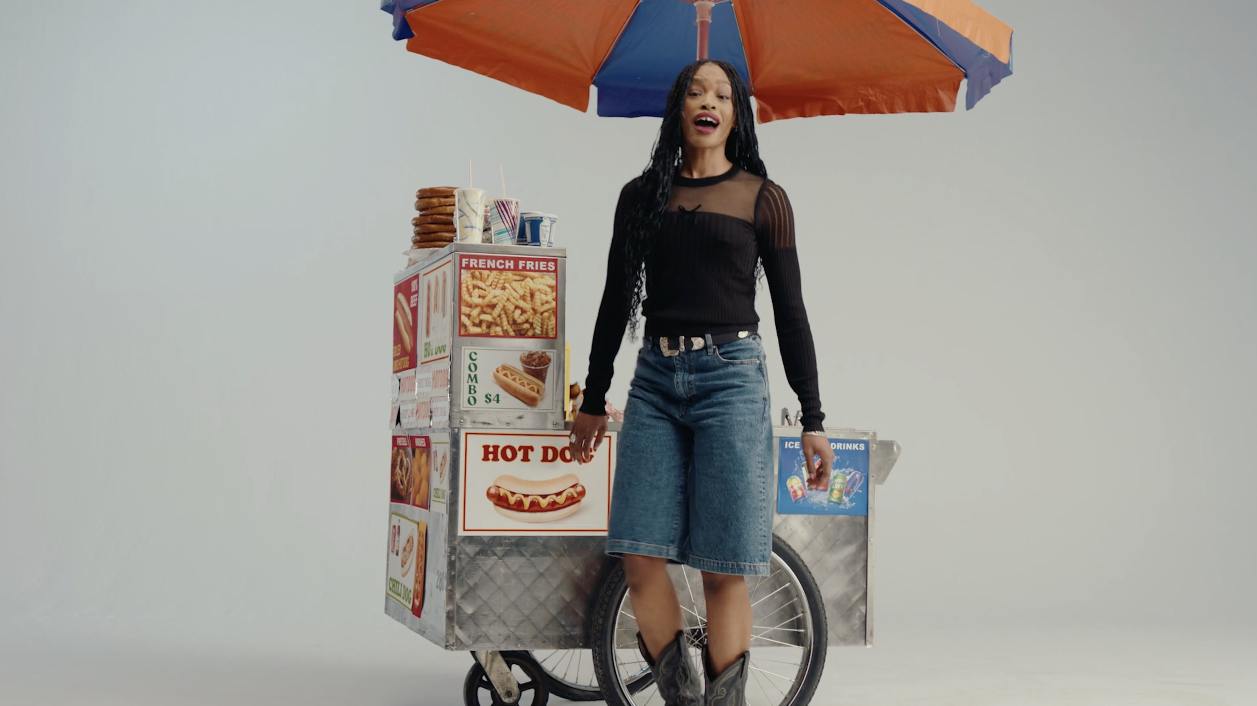 A woman in a black top and blue jeans standing next to a hot dog cart with an orange and blue umbrella, with various food signs and drinks on it, against a plain white background.