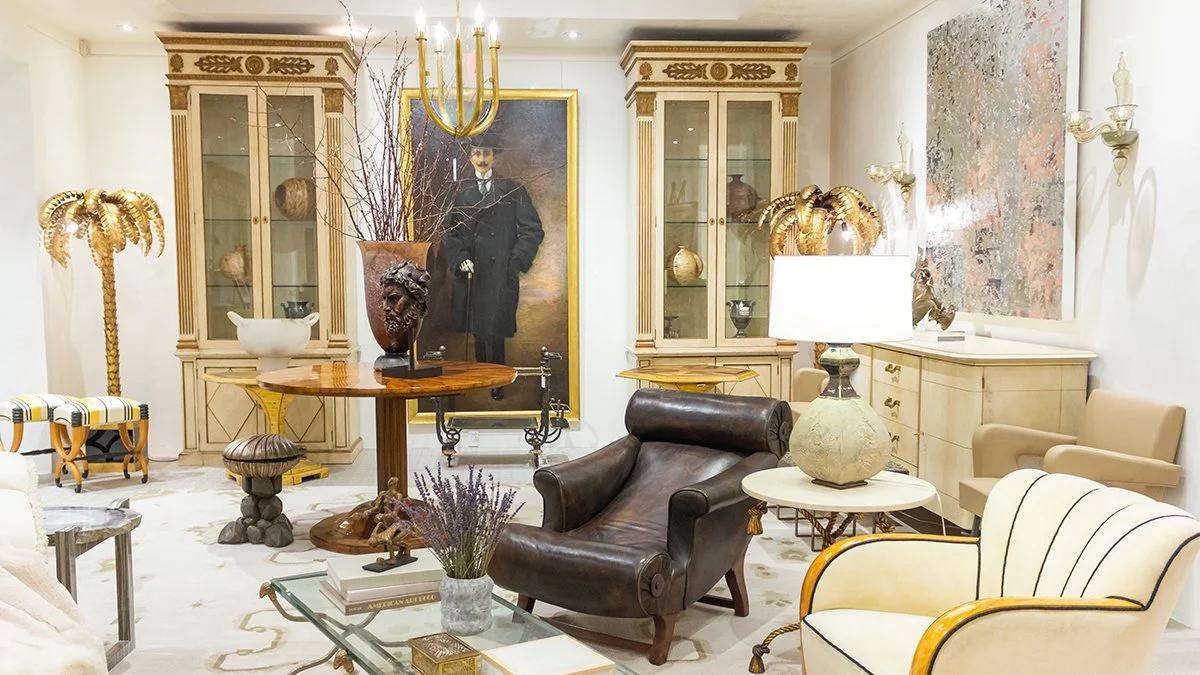 A luxurious living room with antique furniture, including a leather lounge chair, cream armchair, glass coffee table with a vase of lavender, and elegant cabinetry. There are large decorative gold palm trees, an oversized floor lamp, and a large portrait of a man in a suit hanging on the wall. The room features a chandelier and intricate wall sconces, with decorative vases and art pieces.