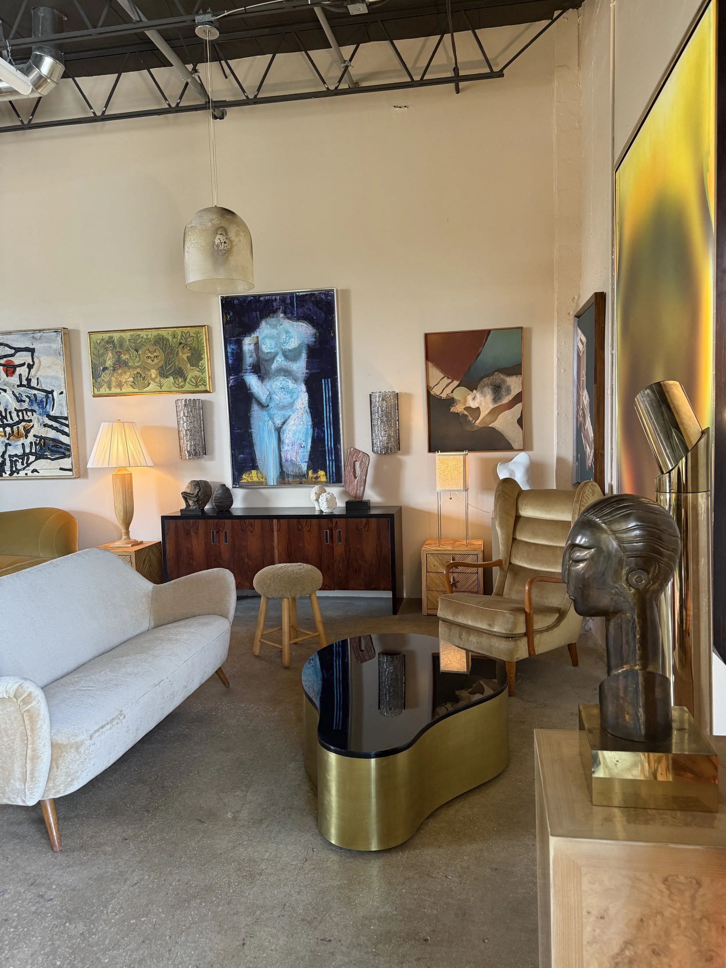 Interior of a living room with various modern and vintage-style furniture, including a white sofa, an ornate armchair, a black and gold coffee table, and art pieces on the wall and on side tables. There are decorative sculptures and lamps, with a high ceiling and exposed industrial-style ductwork.