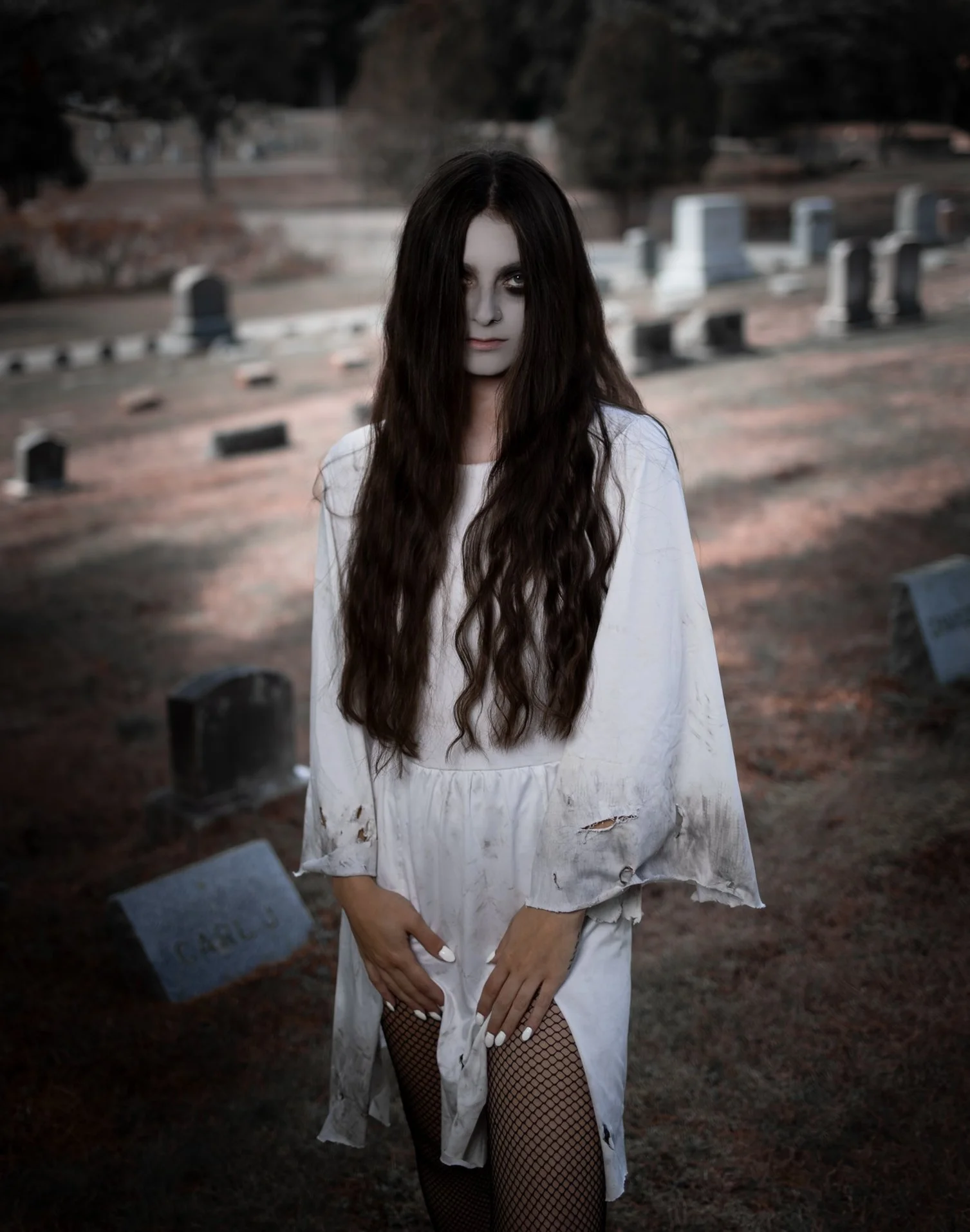 A woman with long dark hair and pale makeup standing in a graveyard, wearing a tattered white dress and fishnet stockings.