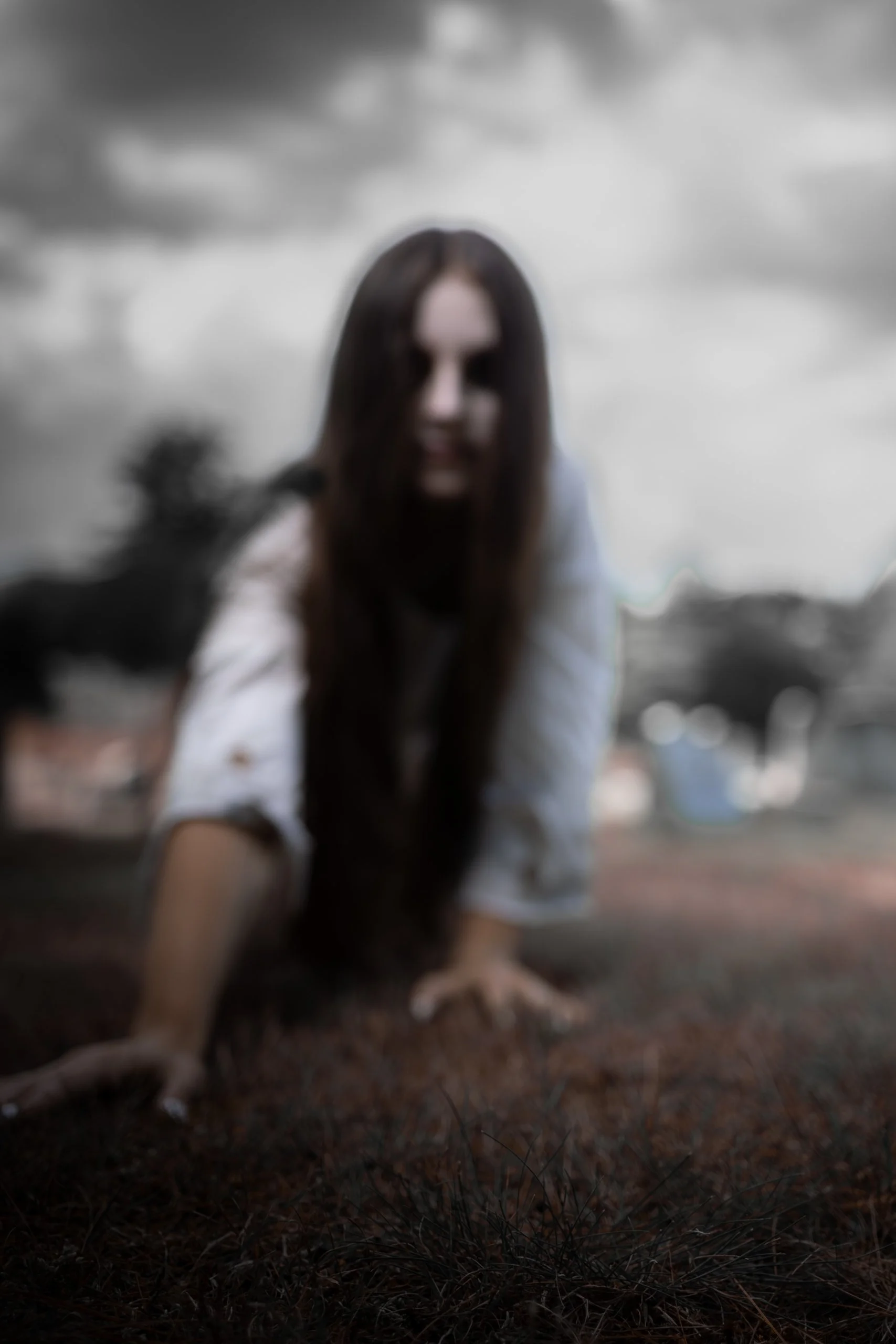 A person with long dark hair crawling on the ground outdoors, with a cloudy sky in the background.