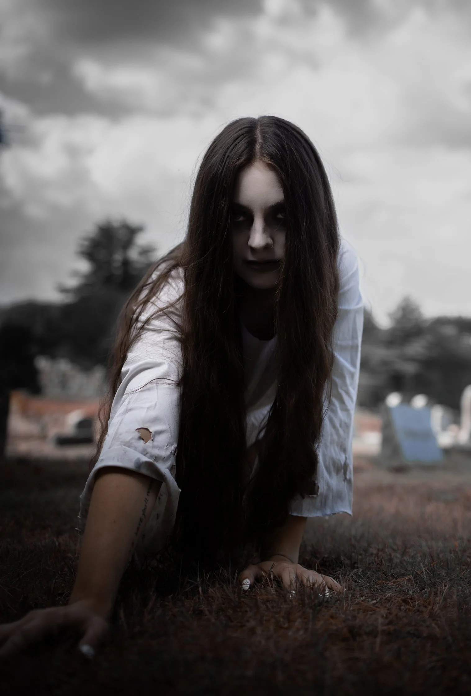 A woman with long dark hair and dark makeup crawling on the ground in a dark, ominous setting with stormy clouds overhead.