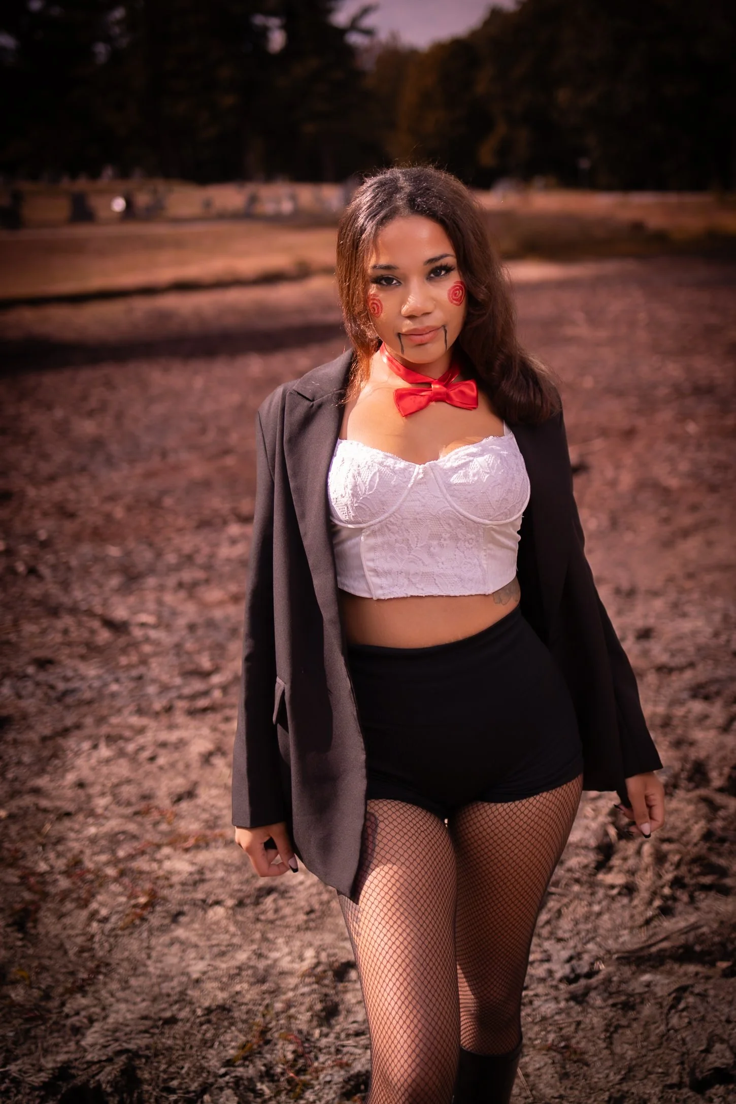 A woman standing outdoors at dusk or night, dressed in a costume with Halloween makeup, fishnet stockings, a white lace crop top, black shorts, a gray blazer, a red bow tie, and face paint resembling a doll or clown with red spirals and lines.