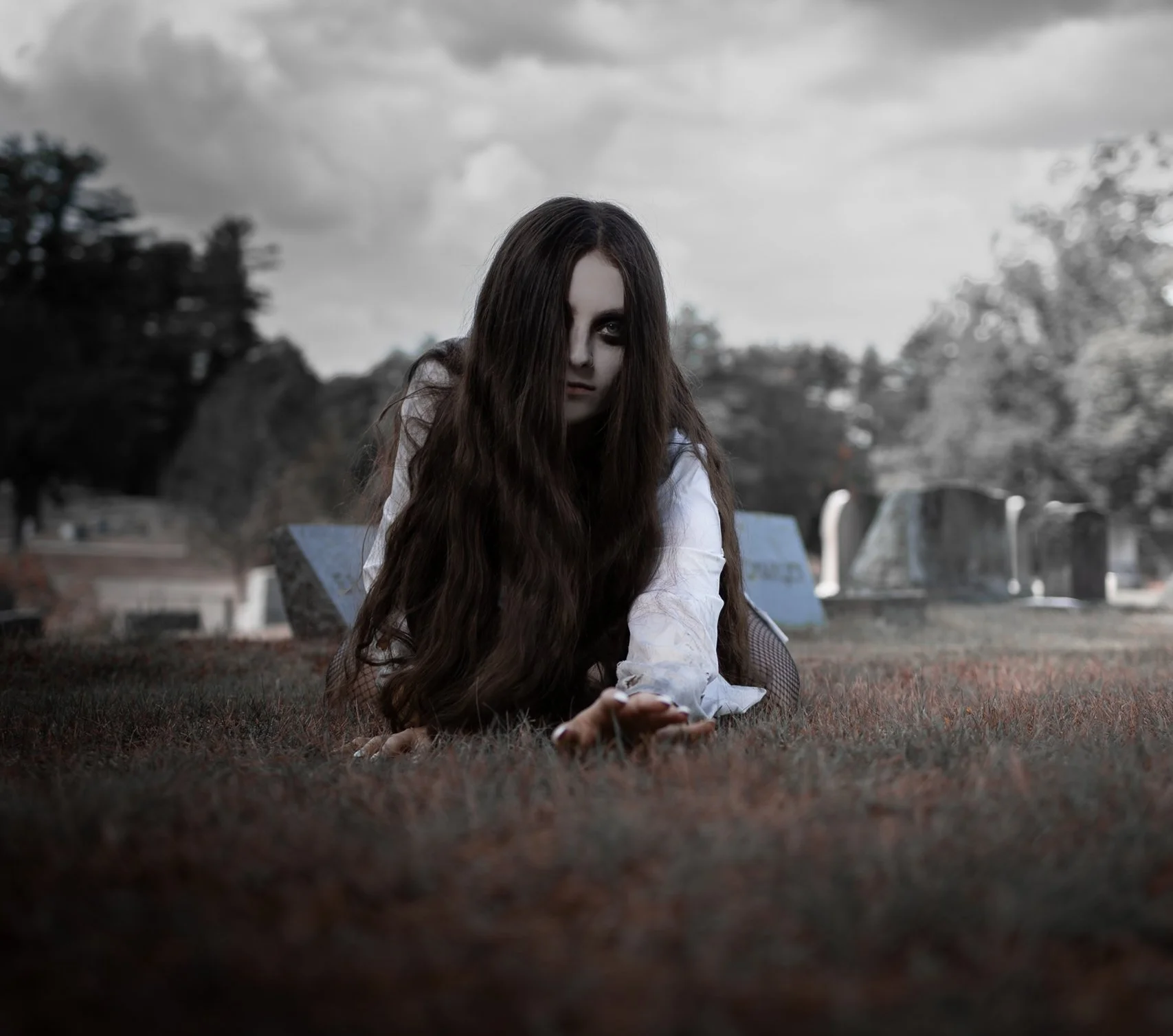 A woman with long dark hair and dark eye makeup is lying on the grass in a cemetery, looking directly at the camera with a somber expression. The background shows blurred tombstones and trees, with a cloudy sky overhead.