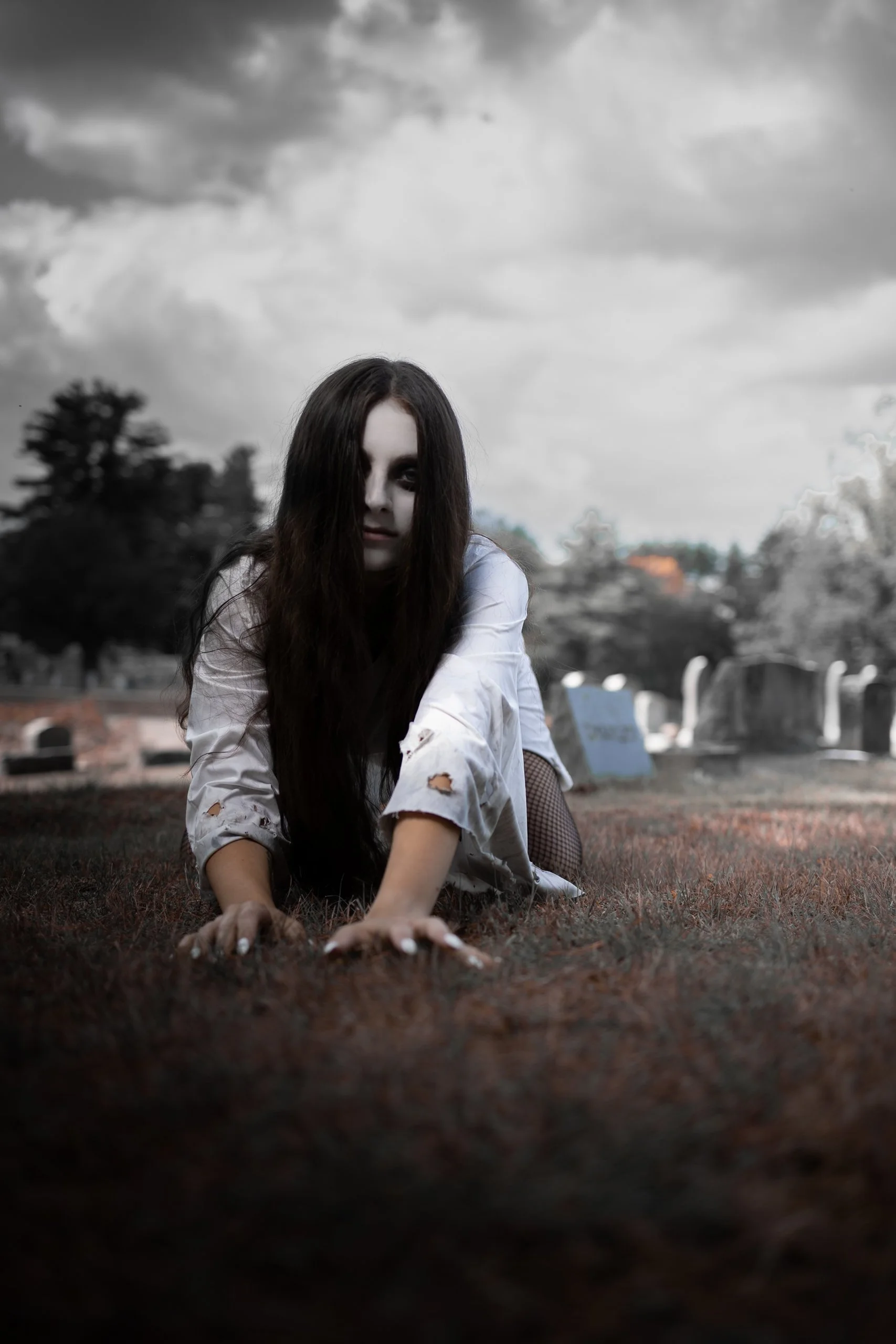 A woman with long dark hair and pale face makeup in a torn white shirt crawling on the ground during daytime in a graveyard.