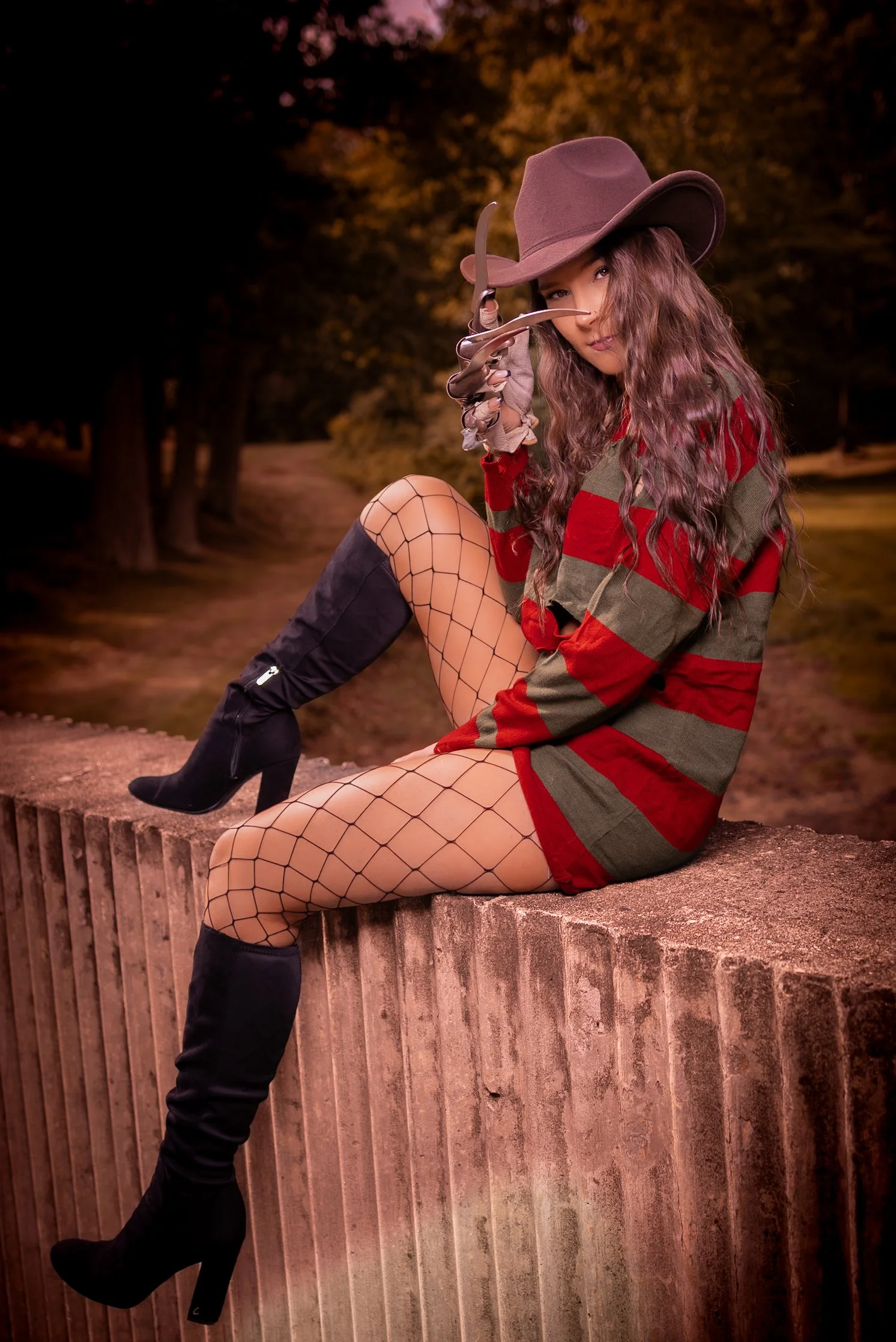A woman dressed as a witch sitting on a brick wall, wearing a striped sweater, fishnet stockings, high-heeled boots, a large witch's hat, and holding a toy sword, with a wooded outdoor background during sunset.