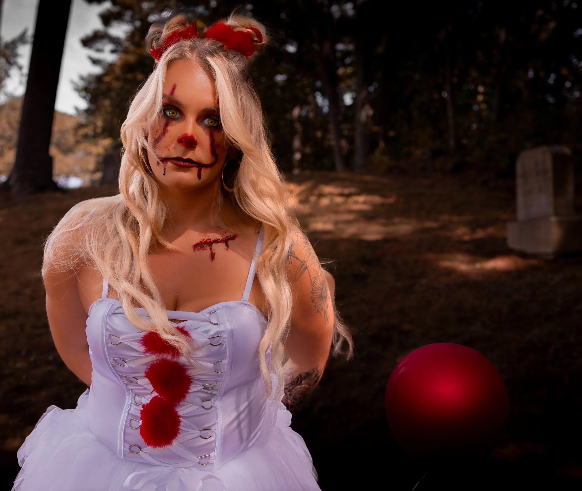 A woman dressed as a creepy clown with blood-like makeup and scars, wearing a white dress with blood stains, standing in a dark outdoor setting with trees and a red balloon nearby.