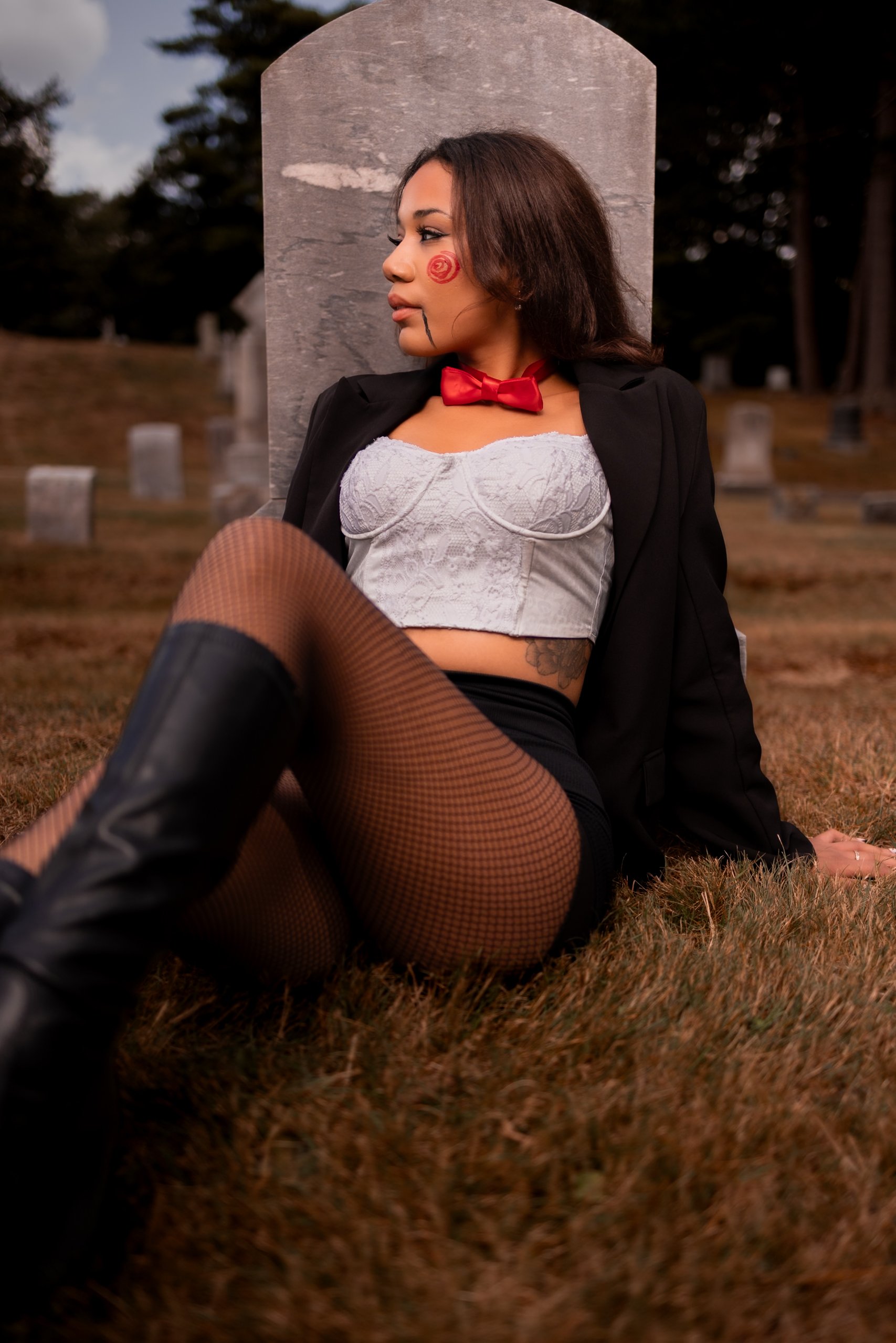 A woman dressed in cosplay attire sitting on the ground in a graveyard, wearing a black blazer, white lace crop top, fishnet stockings, and black boots, with face paint resembling a skull and a red bow tie around her neck.