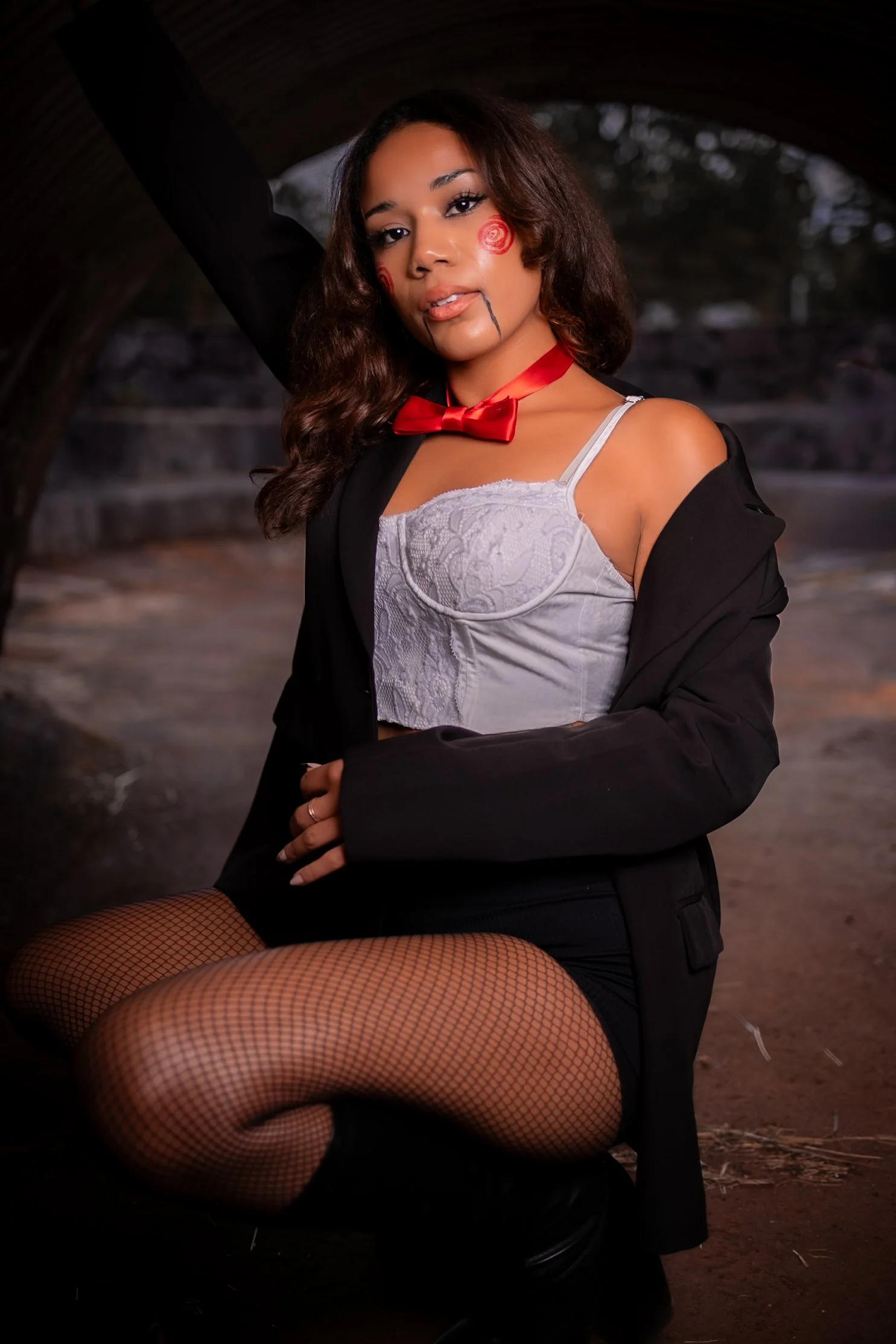 A woman dressed in a costume with face paint, fishnet stockings, a white corset, a black jacket, and a red bow tie, posing under a bridge with one arm raised.